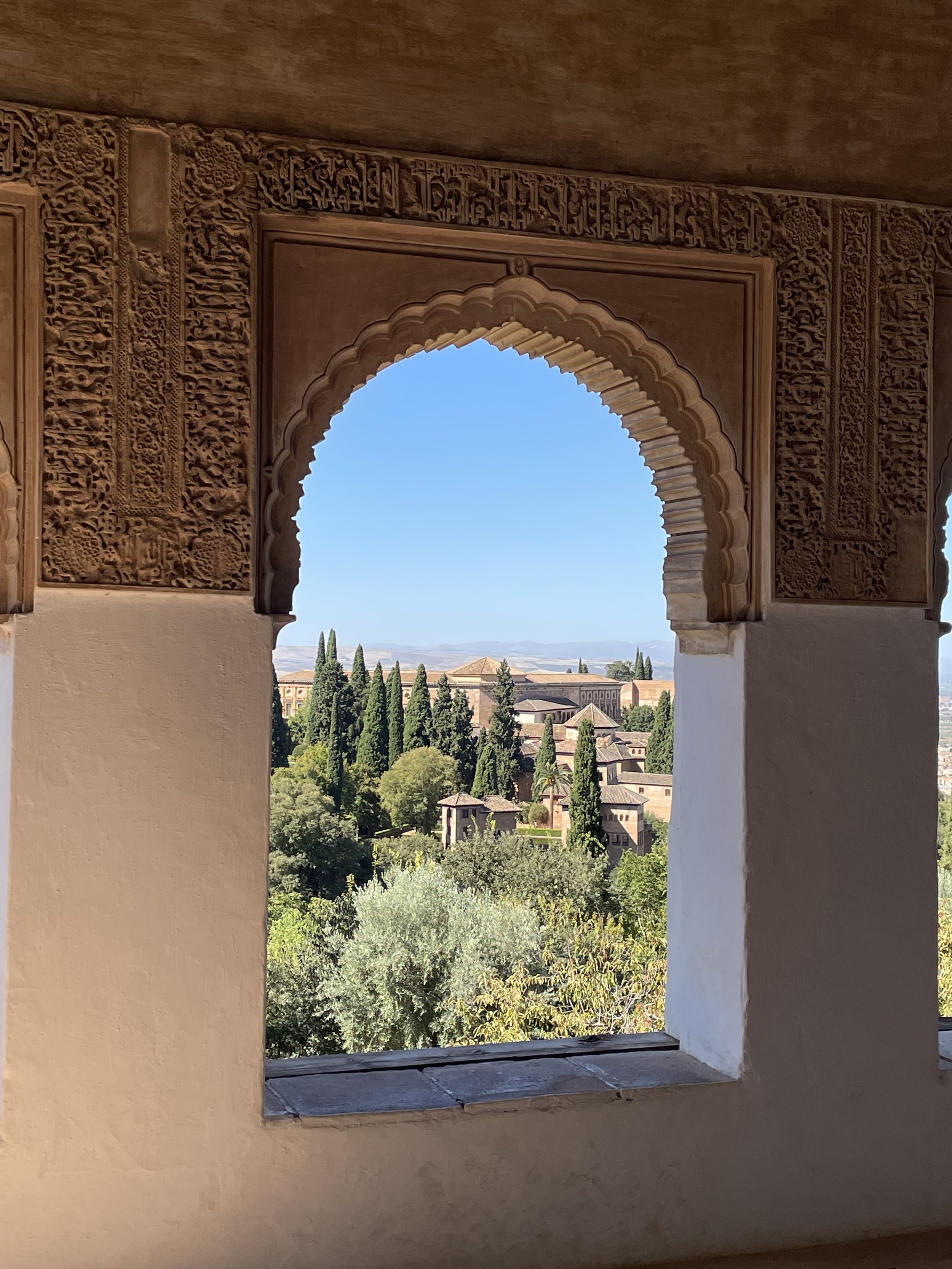 View of a lush green landscape with trees and buildings through a decorative archway in a historical structure, with a clear blue sky in the background.