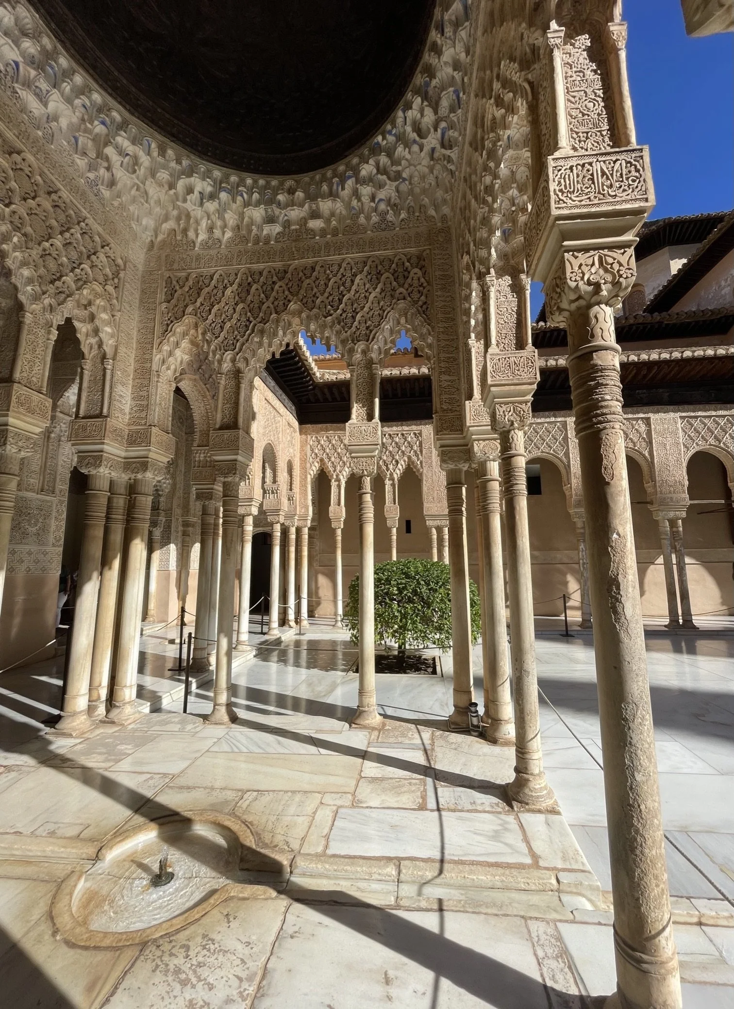 An interior courtyard with ornate marble columns, archways, intricate carvings, and a small fountain with a water spout, all illuminated by natural sunlight.