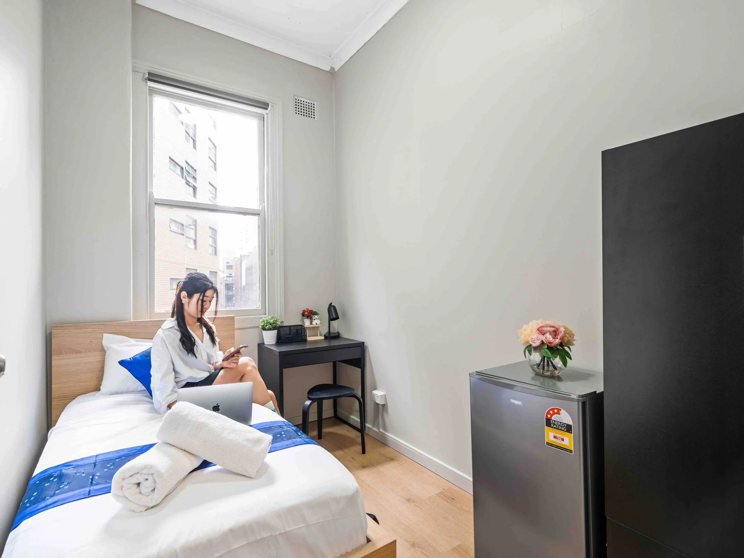 A woman sitting on a bed in a small, minimalist bedroom, looking at her phone. The bed has a rolled towel on it. The room has a window with a view of neighboring buildings, a small black side table with a plant, a lamp, and other items, a mini fridge with a flower on top, and a black wall with a vent.