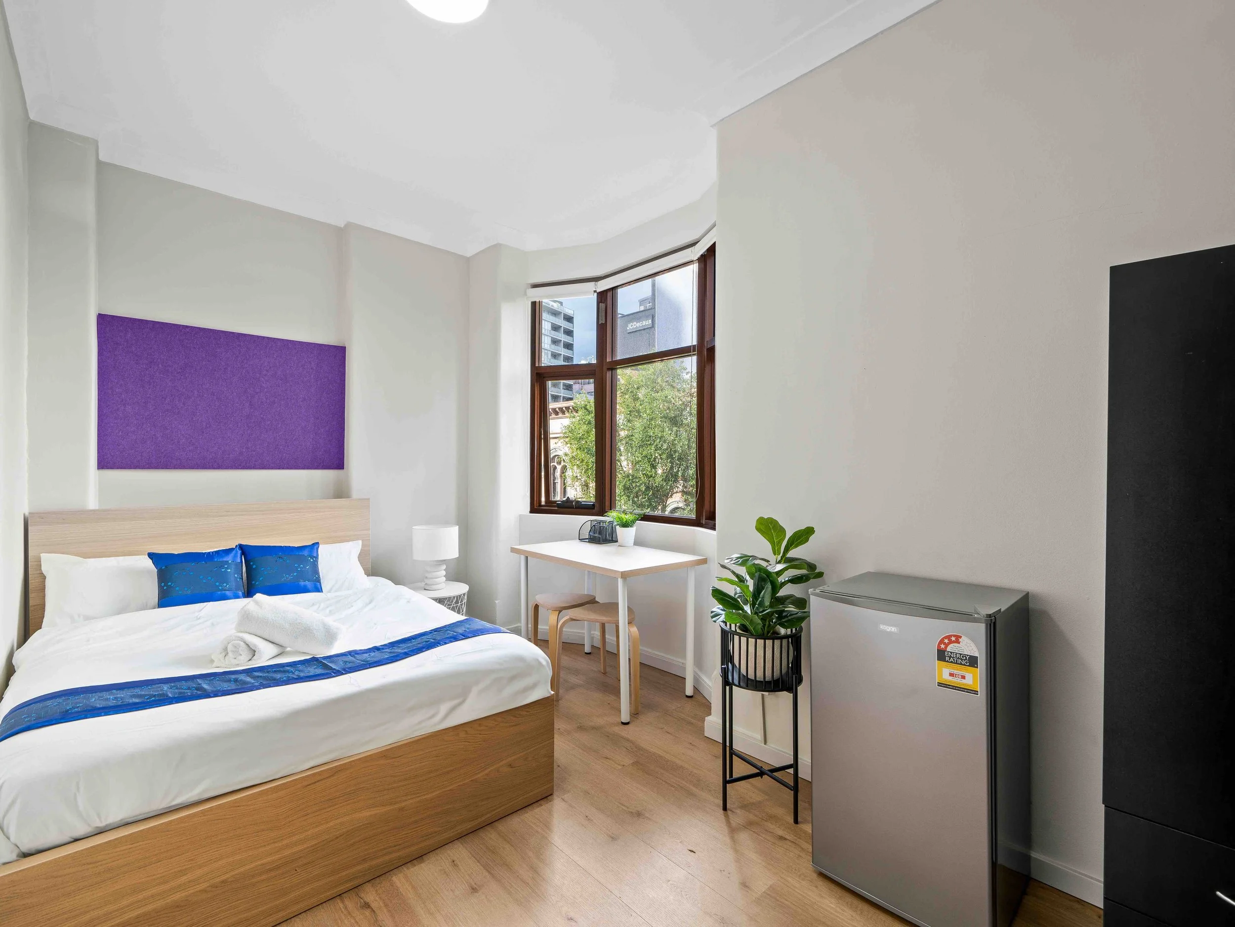 A cozy bedroom with a wooden bed frame, white bedding, and blue pillows. There is a purple wall panel behind the bed, a window with a city view, a small white bedside table with a lamp, a desk with a small potted plant, and a mini fridge or freezer.