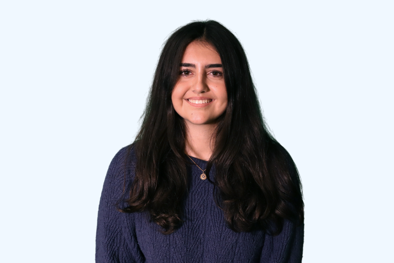 A young woman with long dark hair, wearing a navy blue sweater and a necklace, smiling against a light blue background.