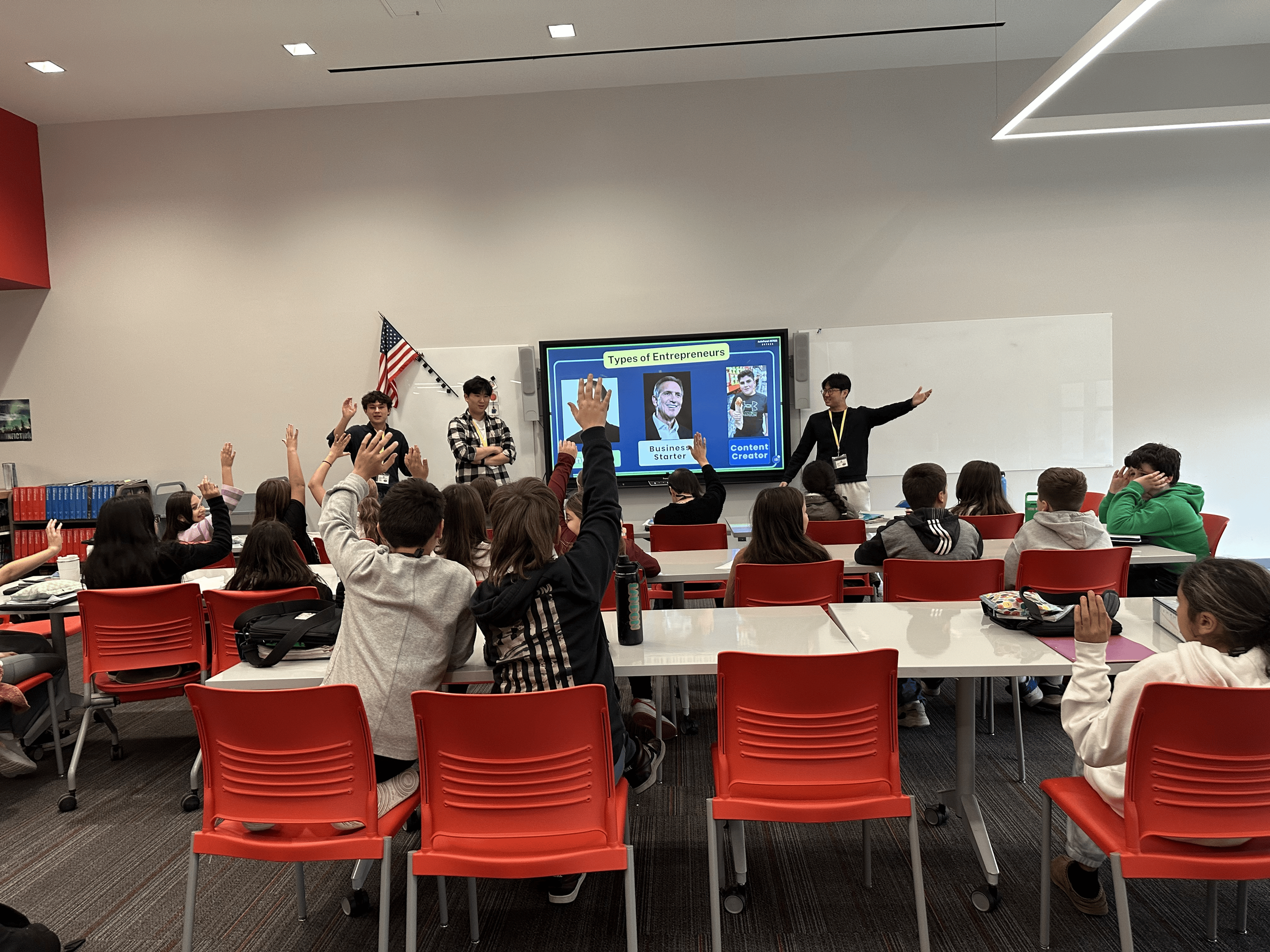 Classroom with students raising hands during lecture on entrepreneurs, with a teacher pointing at a presentation slide.