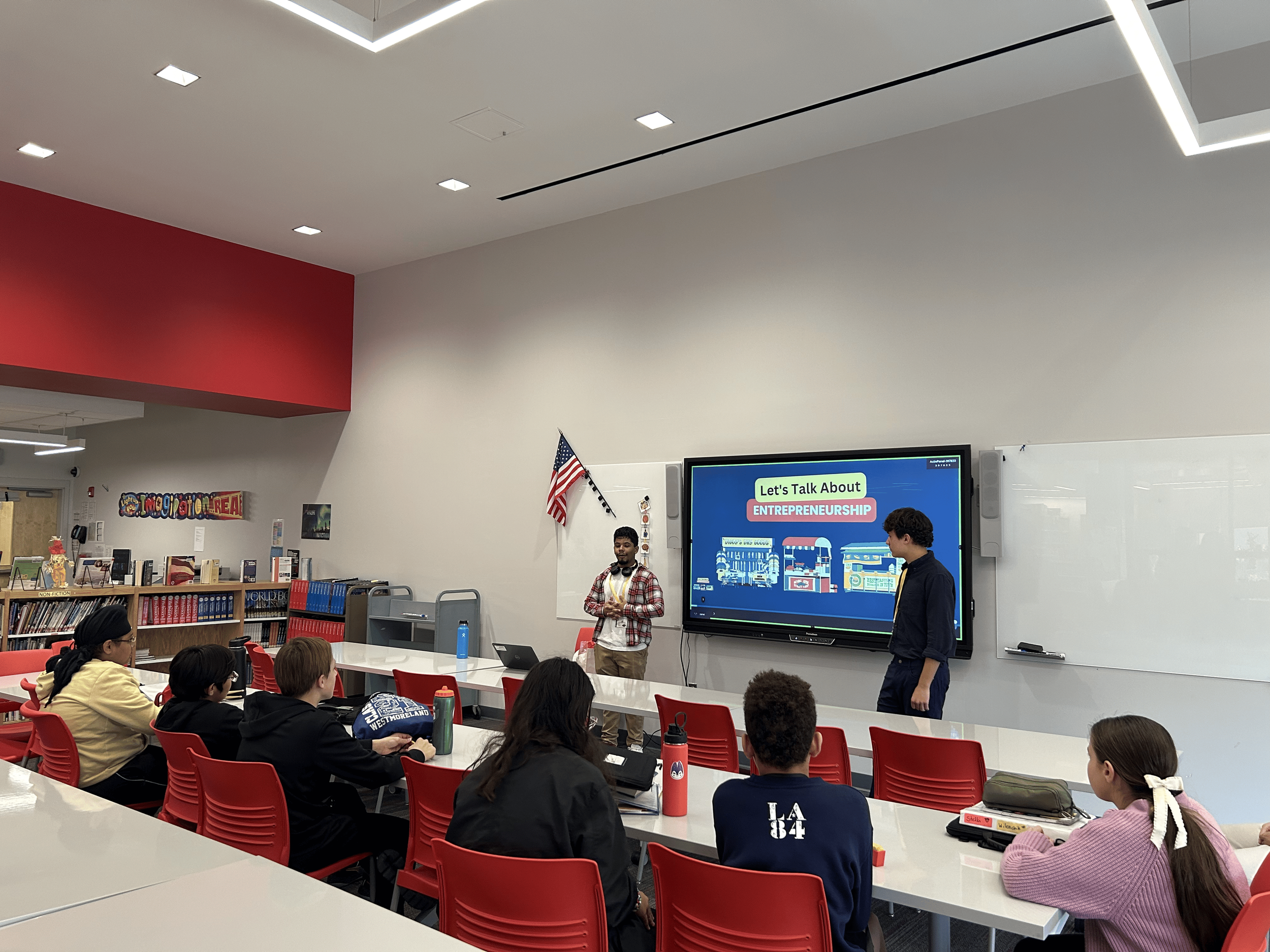 Classroom scene with students sitting at tables, two boys presenting at the front with a large screen displaying 'Let's Talk About ENTREPRENEURSHIP,' American flag on wall, bookshelves on the side.