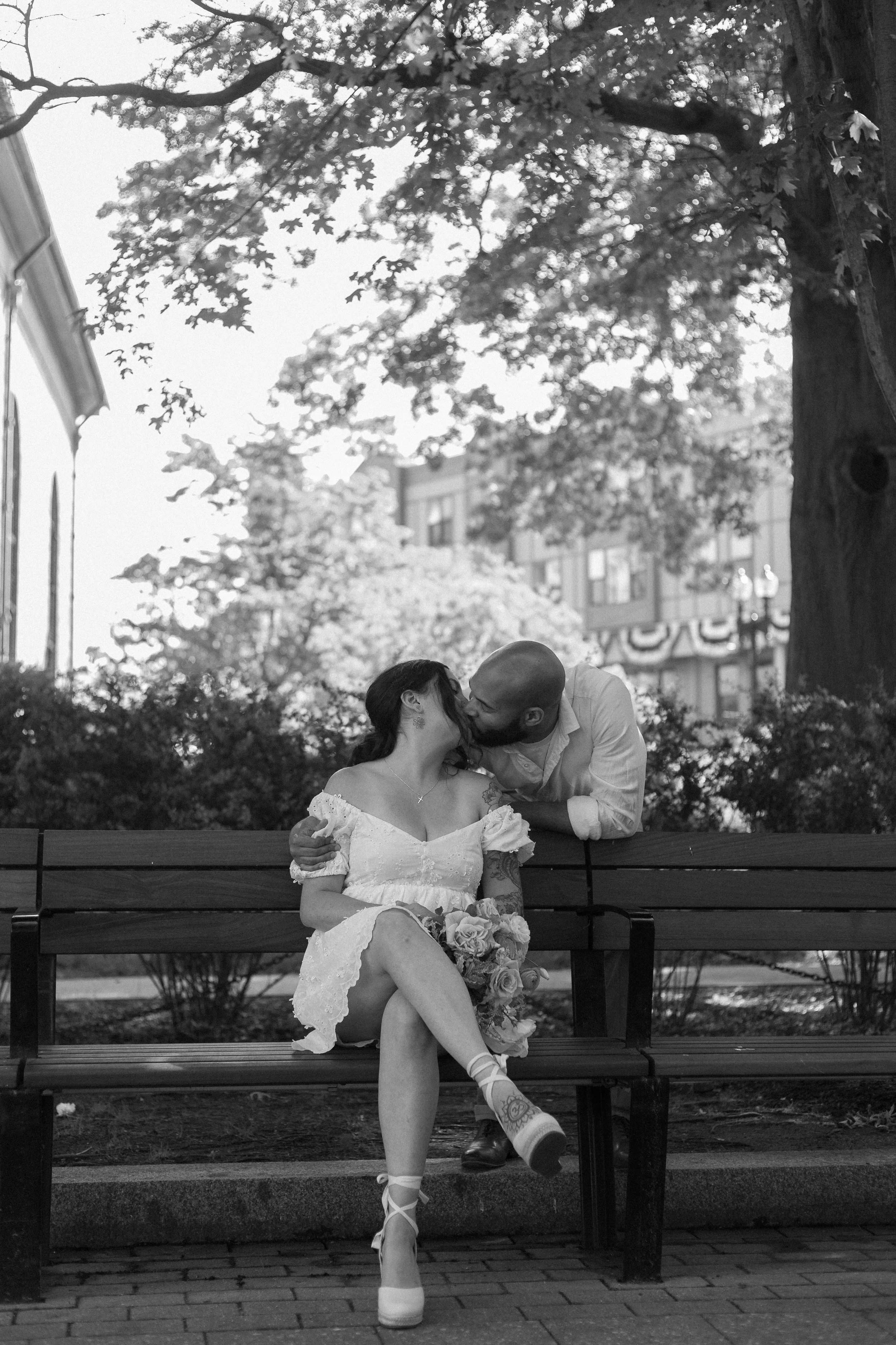A couple sitting on a park bench sharing a kiss, with the woman holding a bouquet of flowers. The woman is wearing a short, white dress with off-the-shoulder sleeves, and the man is leaning over the bench to kiss her, wearing a light-colored shirt. There are trees and buildings in the background.