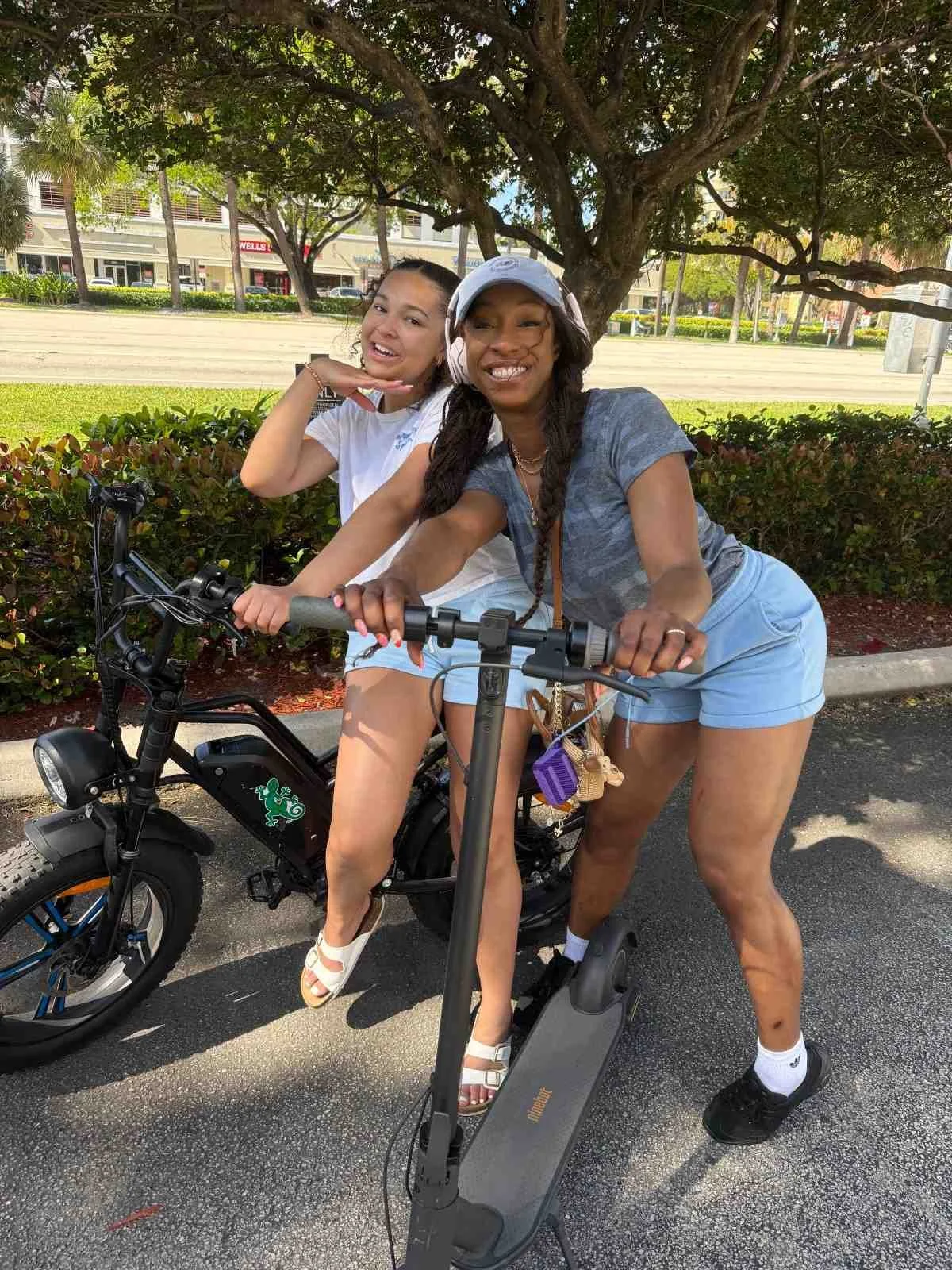 Two women posing with a bicycle and an electric scooter under a tree in an outdoor setting, with shops and greenery in the background.
