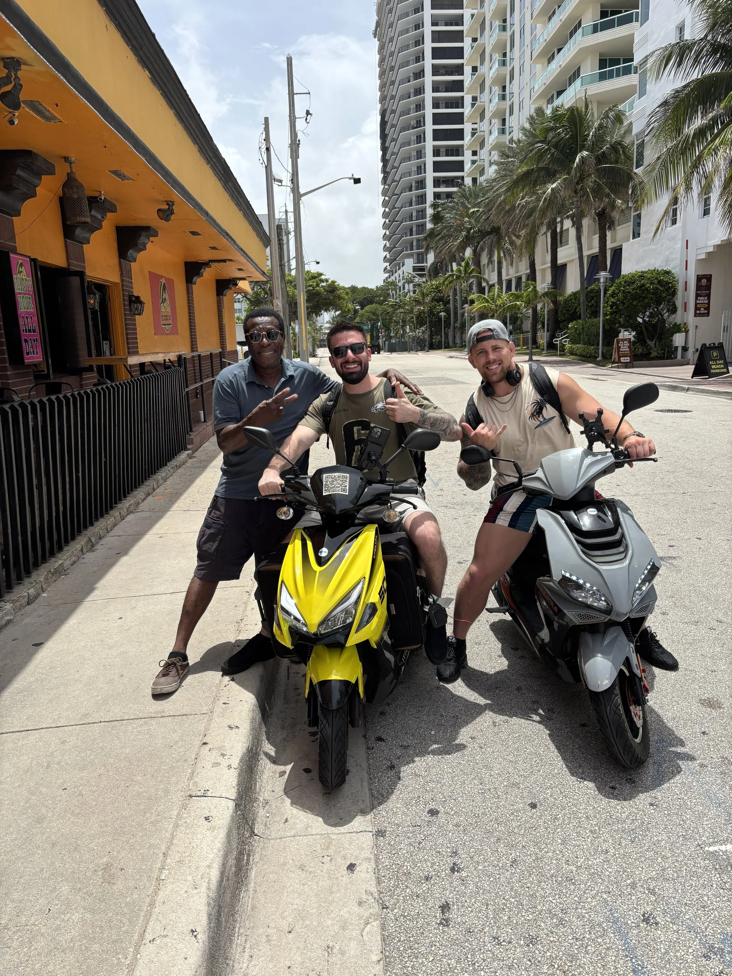 Three men posing with scooters on a city street, with tall buildings, palm trees, and a bright sky in the background.