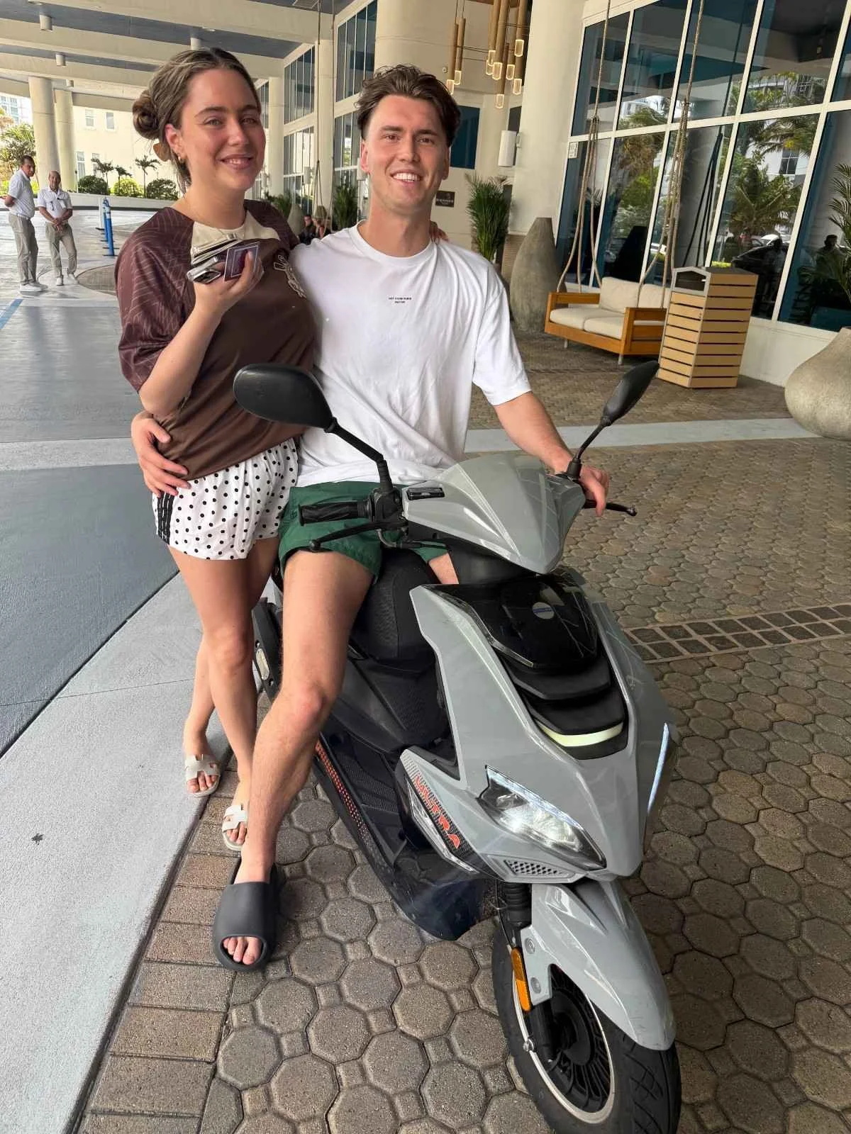 A young woman and a young man are posing together with a scooter outside a building. The woman is wearing a brown shirt and white polka dot shorts, holding a phone, and has her arm around the man. The man is sitting on the scooter, wearing a white t-