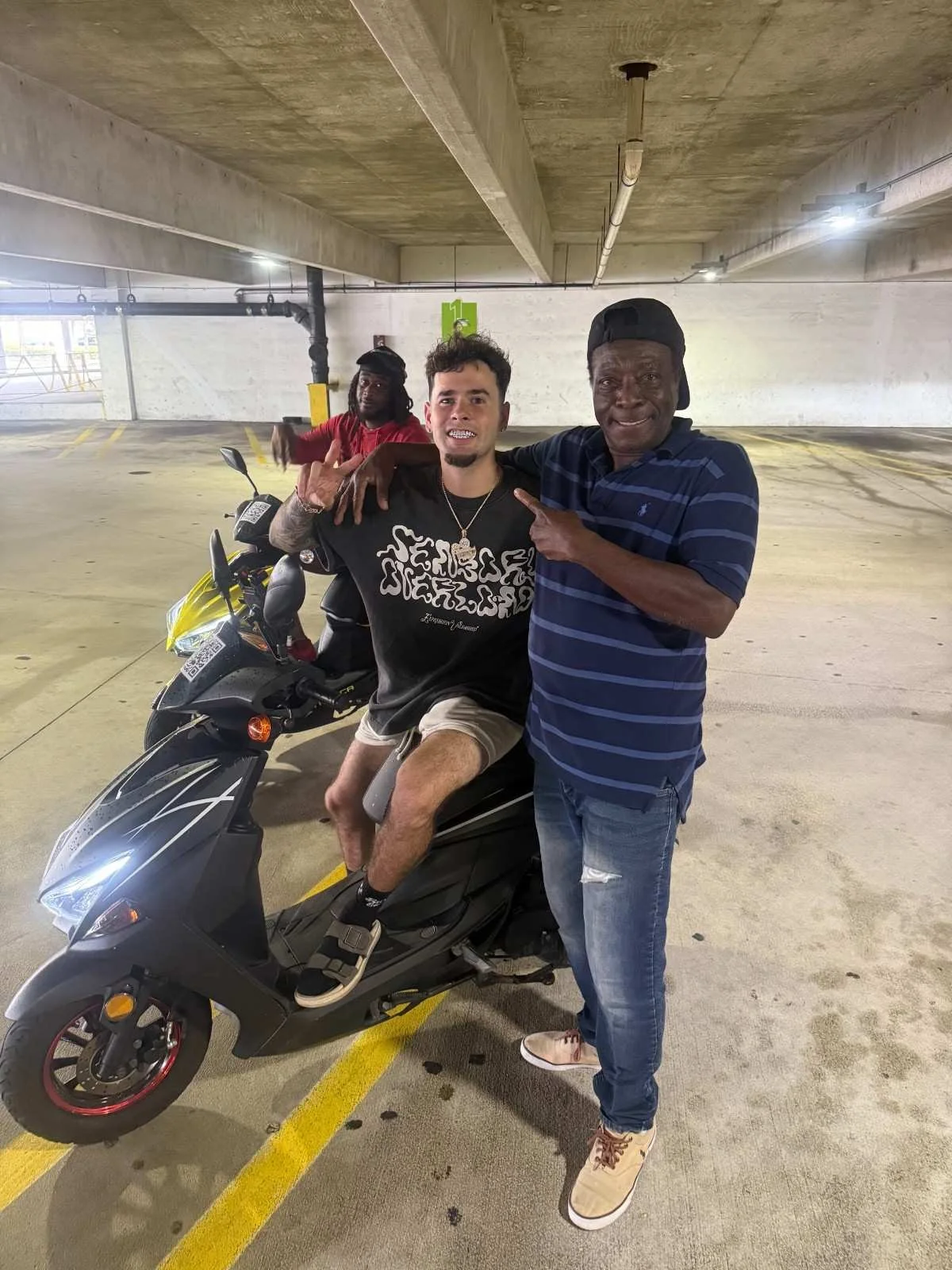 Three men in a parking garage, one sitting on a black scooter and two standing beside him, smiling and posing for the camera.