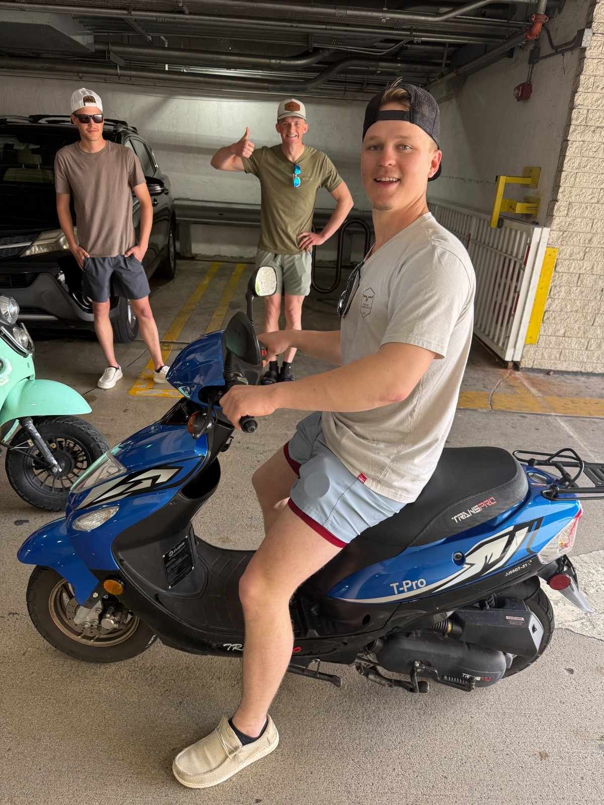 Three young men in casual attire, with sunglasses and hats, are in an indoor parking garage. One is sitting on a blue motor scooter while two others stand behind him, smiling and giving thumbs-up.