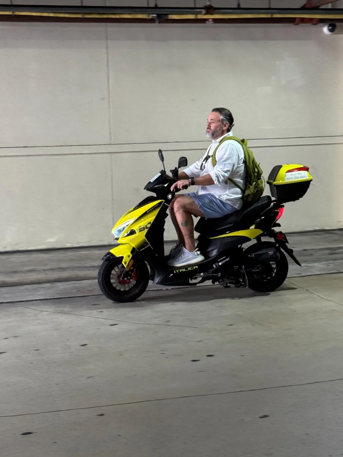 A man riding a yellow and black scooter in an underground tunnel or parking garage, wearing a white shirt, shorts, sneakers, and carrying a green backpack.