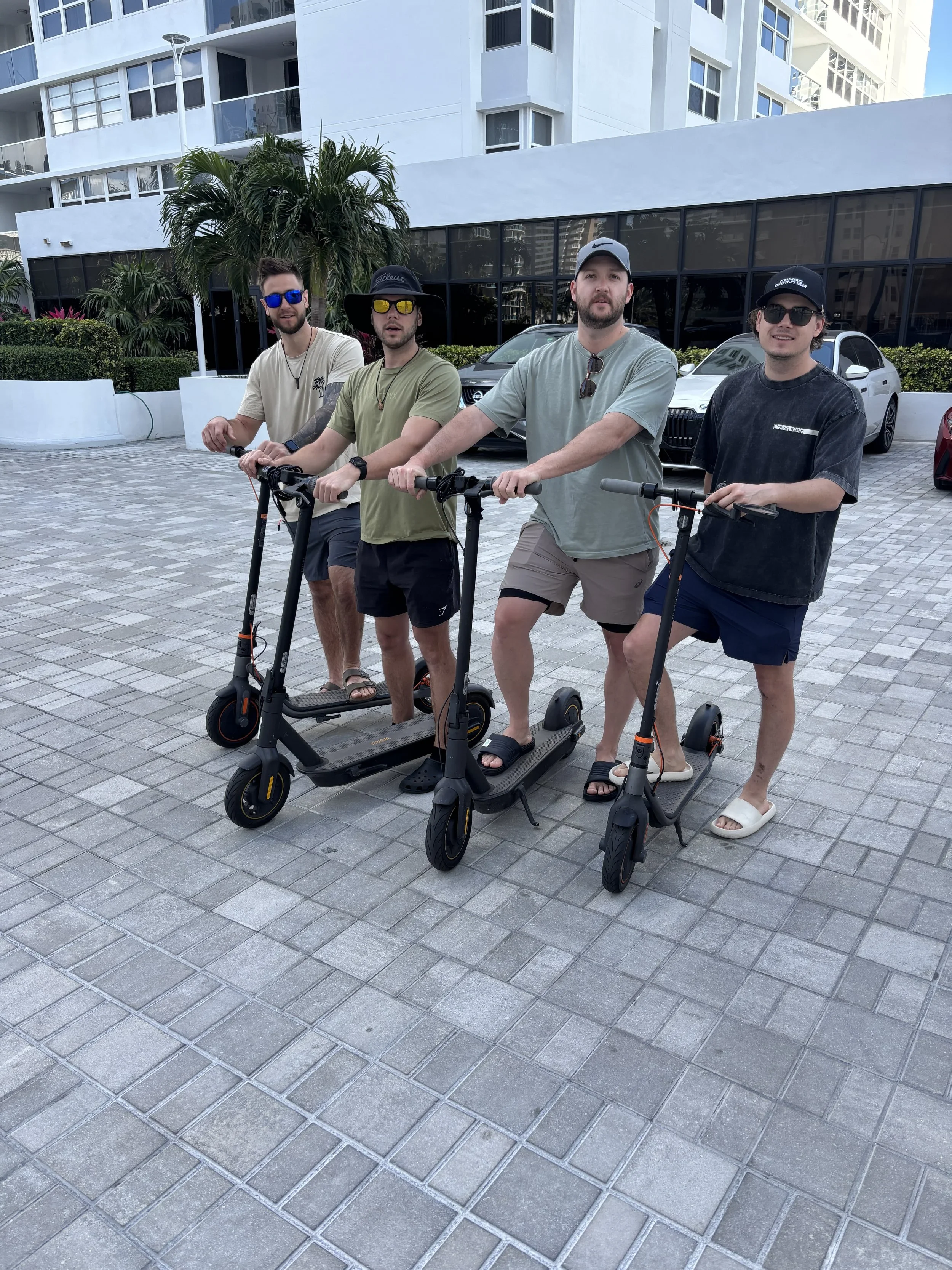 Four men standing on electric scooters outdoors in a city area with modern white buildings, palm trees, and parked cars in the background.