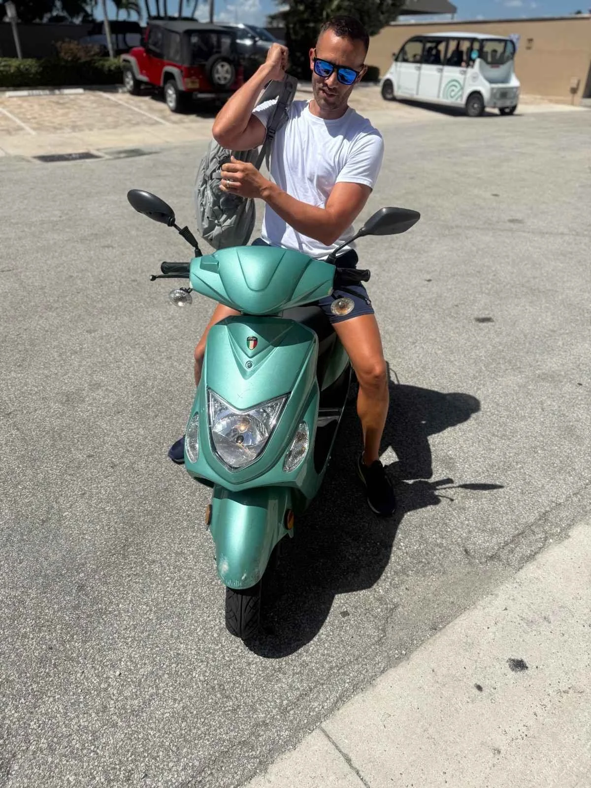 A man wearing sunglasses, a white t-shirt, and shorts sitting on a mint green scooter in a parking lot, with cars in the background.