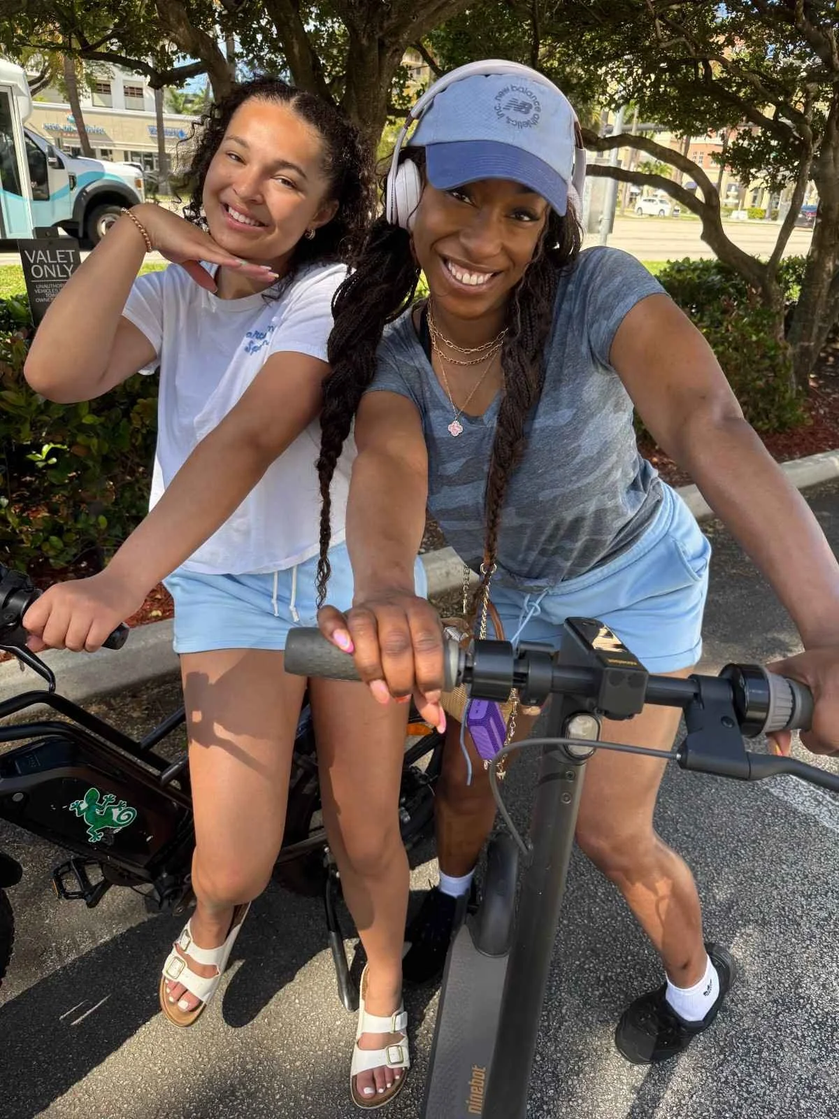 Two women riding electric bikes outdoors, smiling at the camera. One woman is wearing a dark gray shirt, light blue shorts, a blue cap, and headphones. The other woman is wearing a white T-shirt, light blue shorts, and sandals.