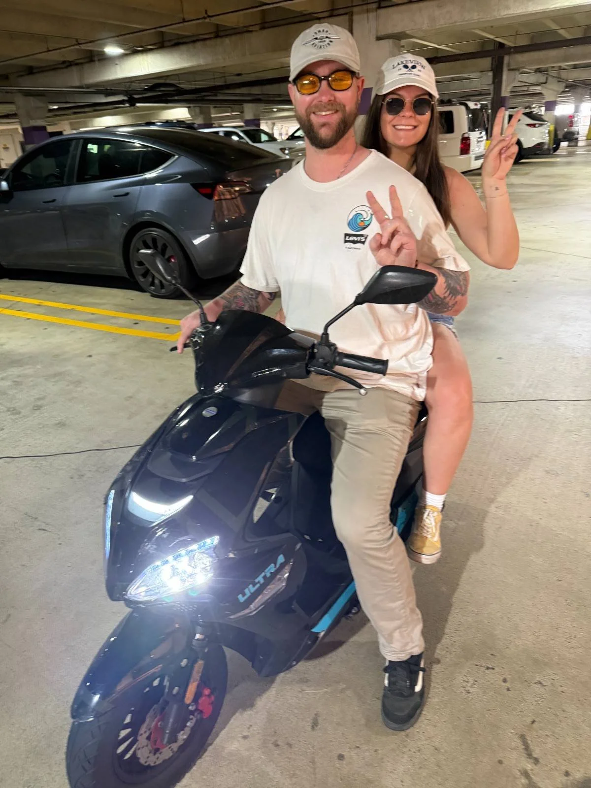 A man and woman sitting on a black electric scooter in a parking garage. The man is wearing a white cap, sunglasses, a white t-shirt with a wave logo, and beige pants. The woman is behind him, smiling and making a peace sign with her right hand. She 