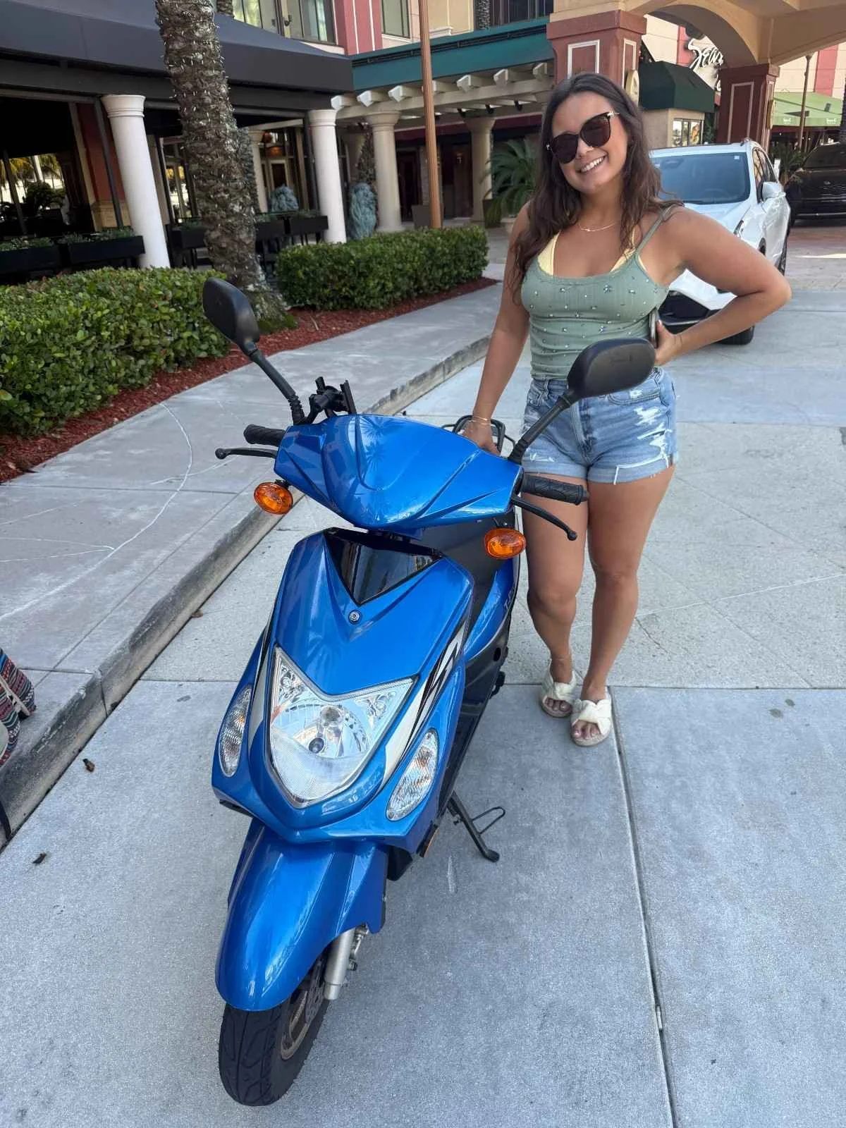A woman wearing sunglasses, a green tank top, and denim shorts standing next to a blue motorcycle in a parking lot.
