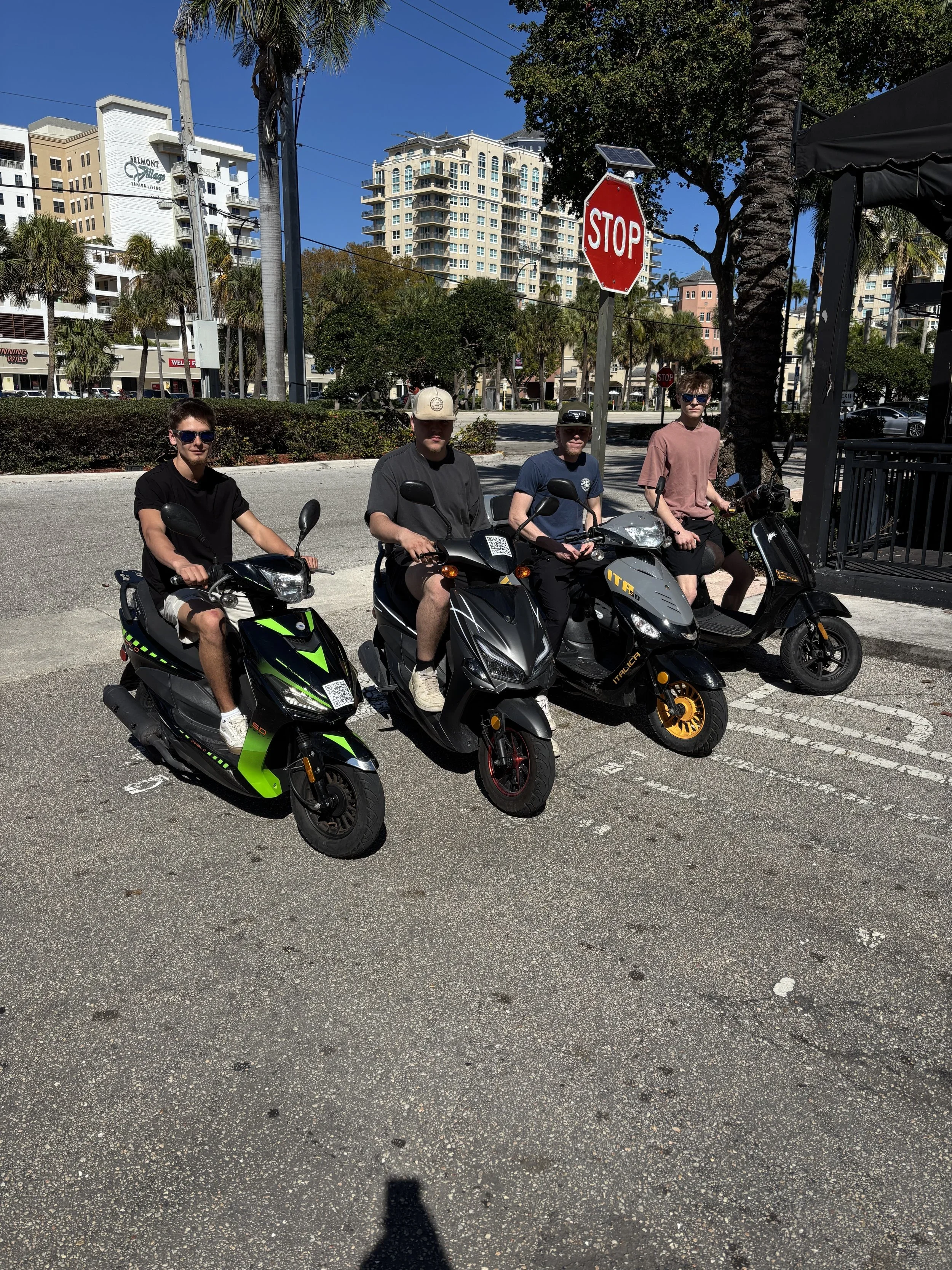 Four individuals on scooters and motorcycles parked on the street, with a background of tall buildings, palm trees, and a stop sign under a clear blue sky.