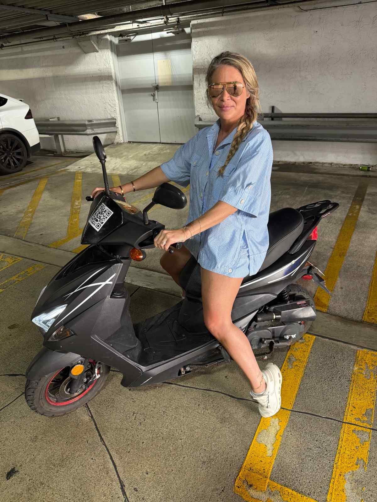 A woman wearing sunglasses and a blue striped shirt is sitting on a black scooter in a parking garage.