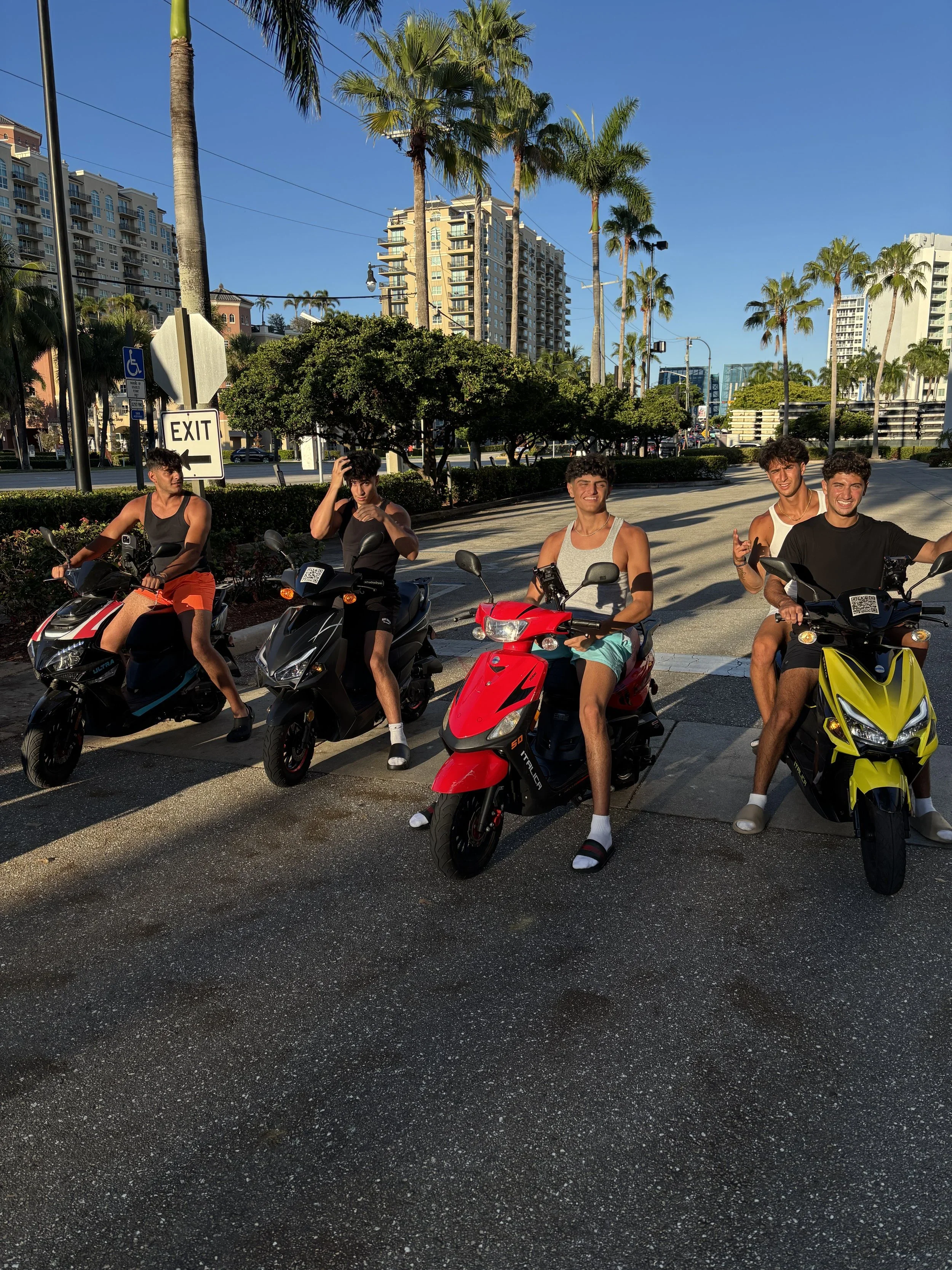 Five young men on scooters lined up on a city street with tall buildings, palm trees, and a clear blue sky in the background.