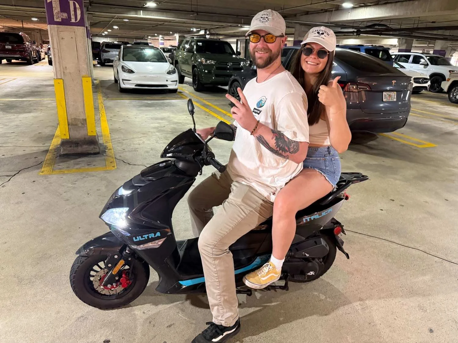 A man and a woman sitting on a black electric scooter in an indoor parking garage. The man is wearing a white cap, sunglasses, a white t-shirt, and khaki pants, making a peace sign. The woman is wearing a beige crop top, denim shorts, and sunglasses,