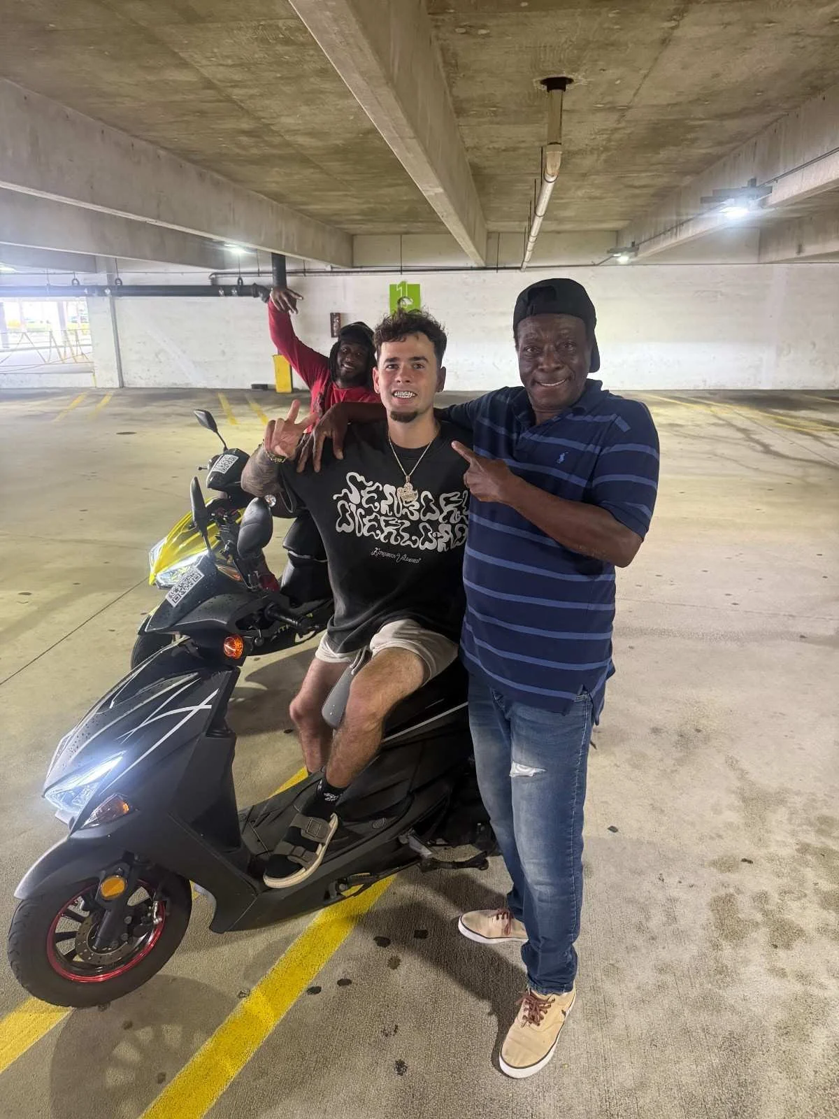 Three men posing in a parking garage, with a motorcycle. One man sitting on the motorcycle, two men standing beside it, making hand gestures.