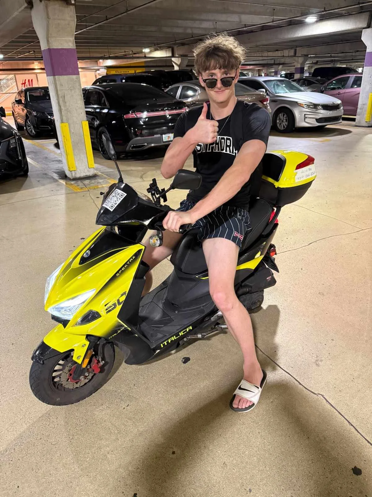 Young man sitting on a yellow and black electric scooter in a parking garage, giving a thumbs-up and smiling. He is wearing sunglasses, a black t-shirt, striped shorts, and slides.
