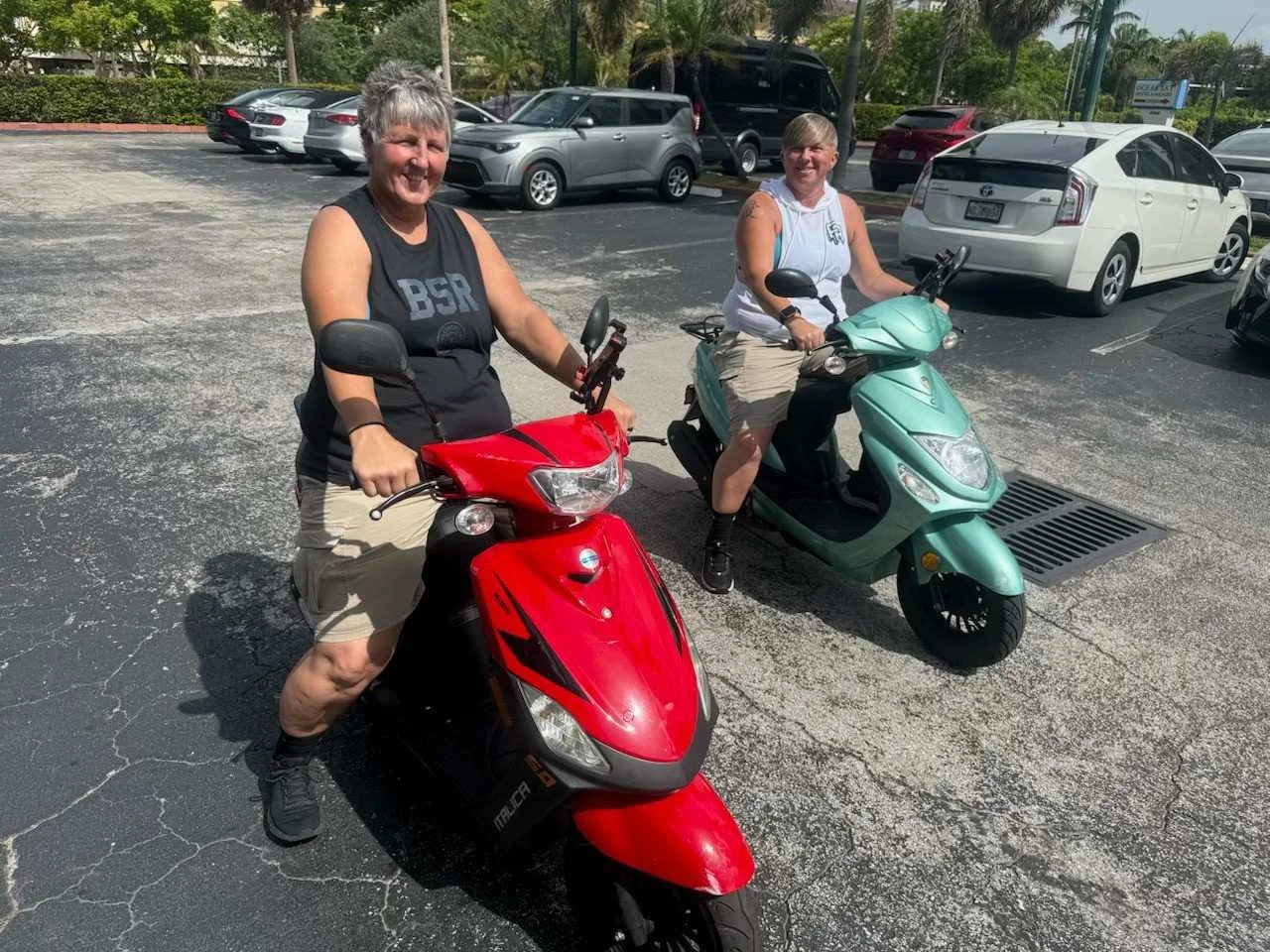 Two women with short gray hair sitting on scooters in a parking lot, smiling at the camera. One woman is on a red scooter, and the other on a green scooter. There are several parked cars in the background and trees around the lot.