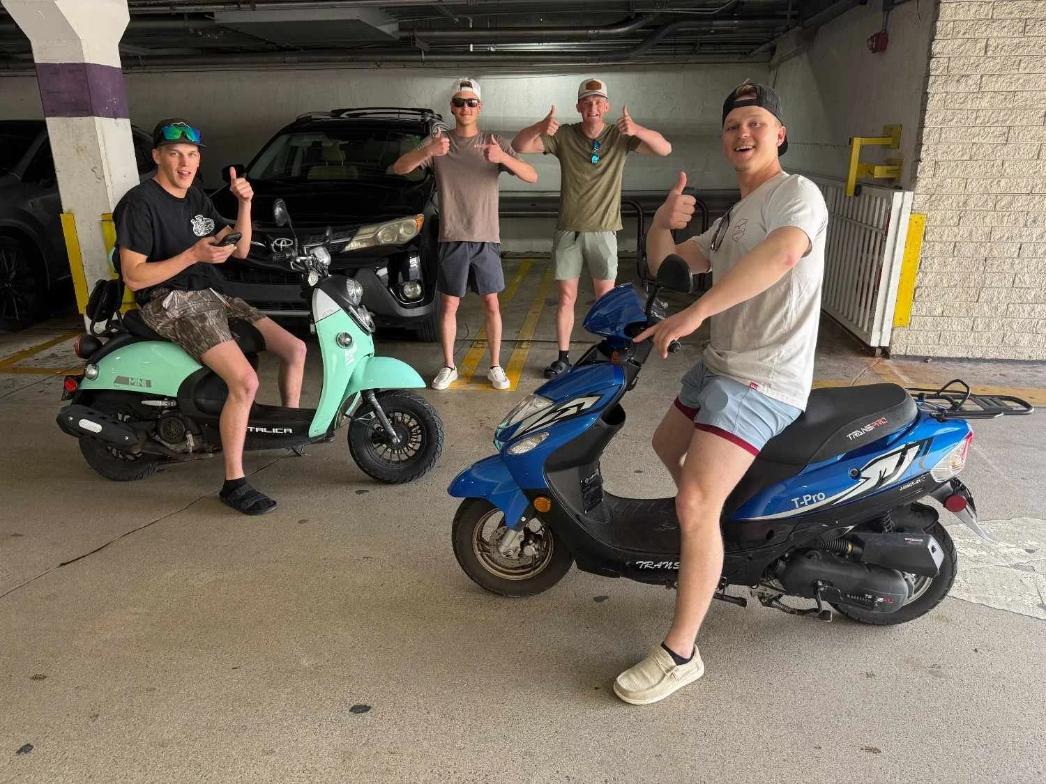 Four young men in casual summer clothing are in an indoor parking garage, with two scooters and a black car in the background. Two are seated on scooters, and two are standing behind them, smiling and giving thumbs-up.