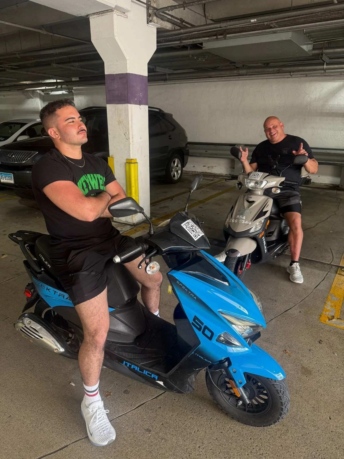 Two men with electric scooters in a parking garage. The man on the left is sitting on a blue scooter with arms crossed, wearing black shorts, white sneakers, and a black T-shirt with green text. The man on the right is sitting on a white scooter, mak