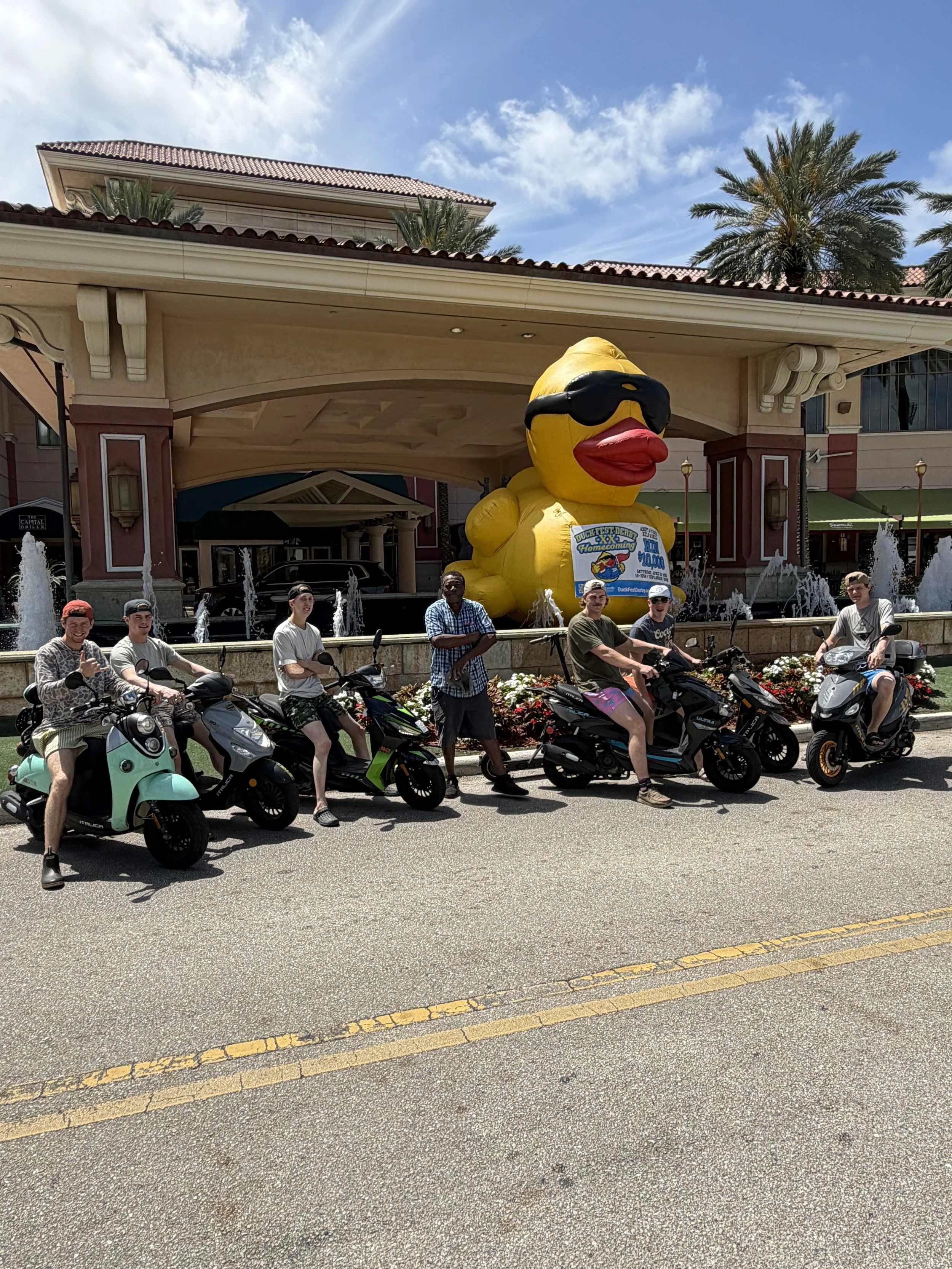Six people on scooters in front of a fountain with a large yellow rubber duck wearing sunglasses and holding a sign, outside a shopping mall under a blue sky with clouds.