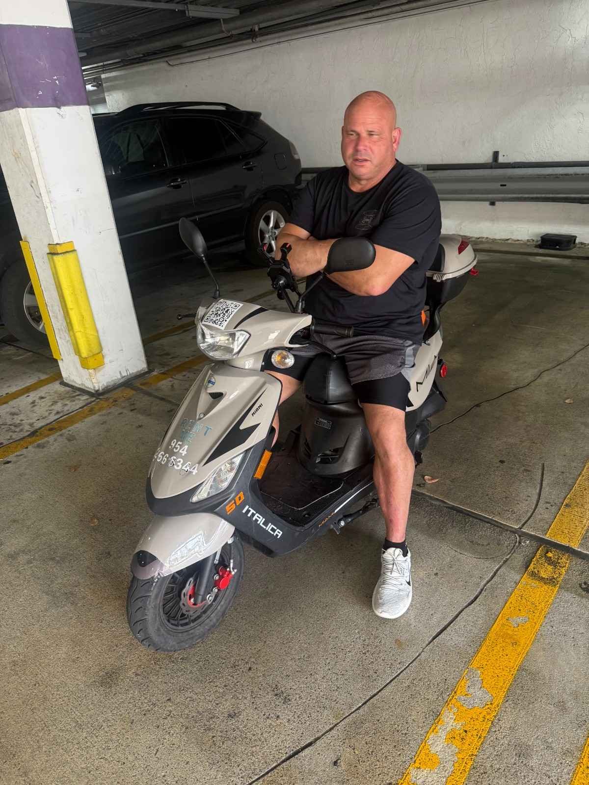 A man sits on a white electric scooter in an underground parking garage, with a black SUV parked behind him and concrete walls around.