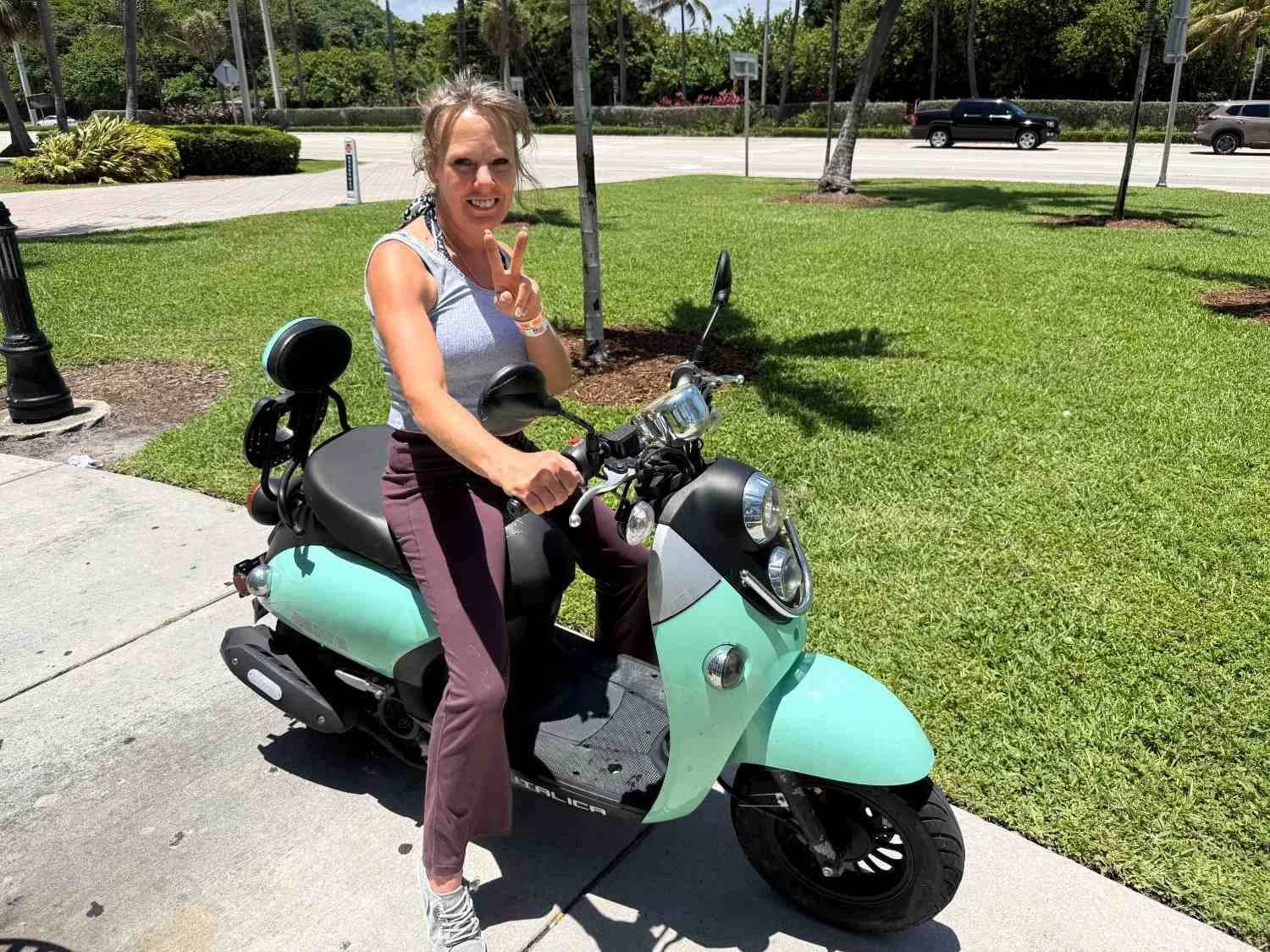 A woman sitting on a light blue scooter, making a peace sign with her right hand, outdoors on a sunny day.