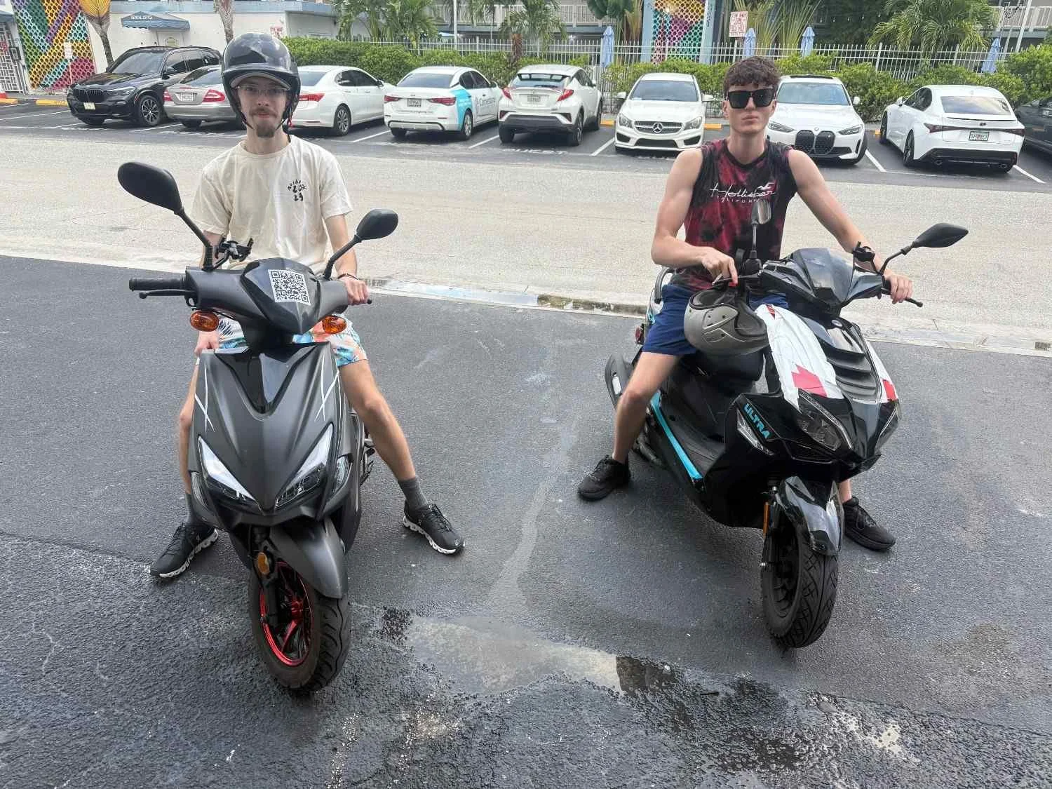 Two young men sitting on black scooters on a parking lot, with cars in the background.