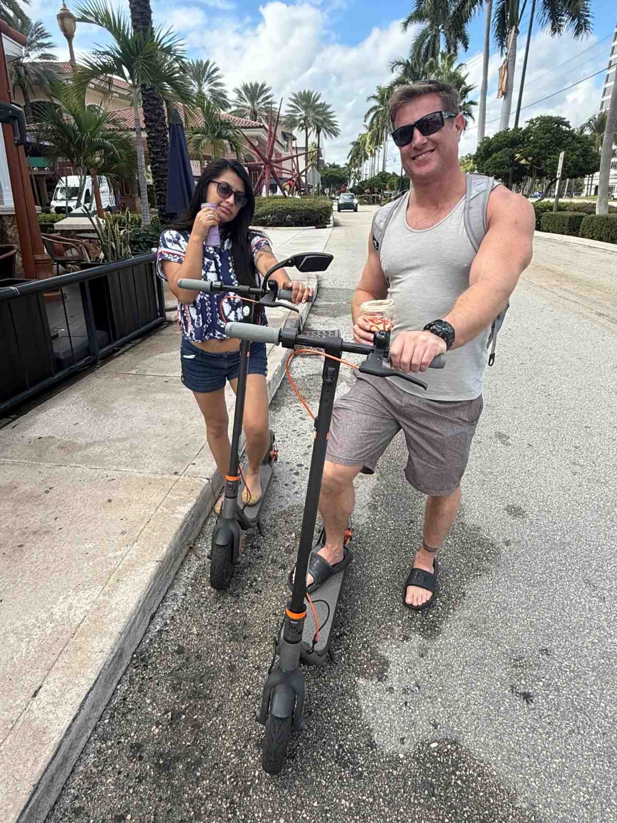 A man and a woman standing on a sidewalk with electric scooters, palm trees, and buildings in the background. The man is smiling, wearing sunglasses, a gray sleeveless shirt, shorts, and sandals, holding a drink. The woman has long dark hair, sunglas