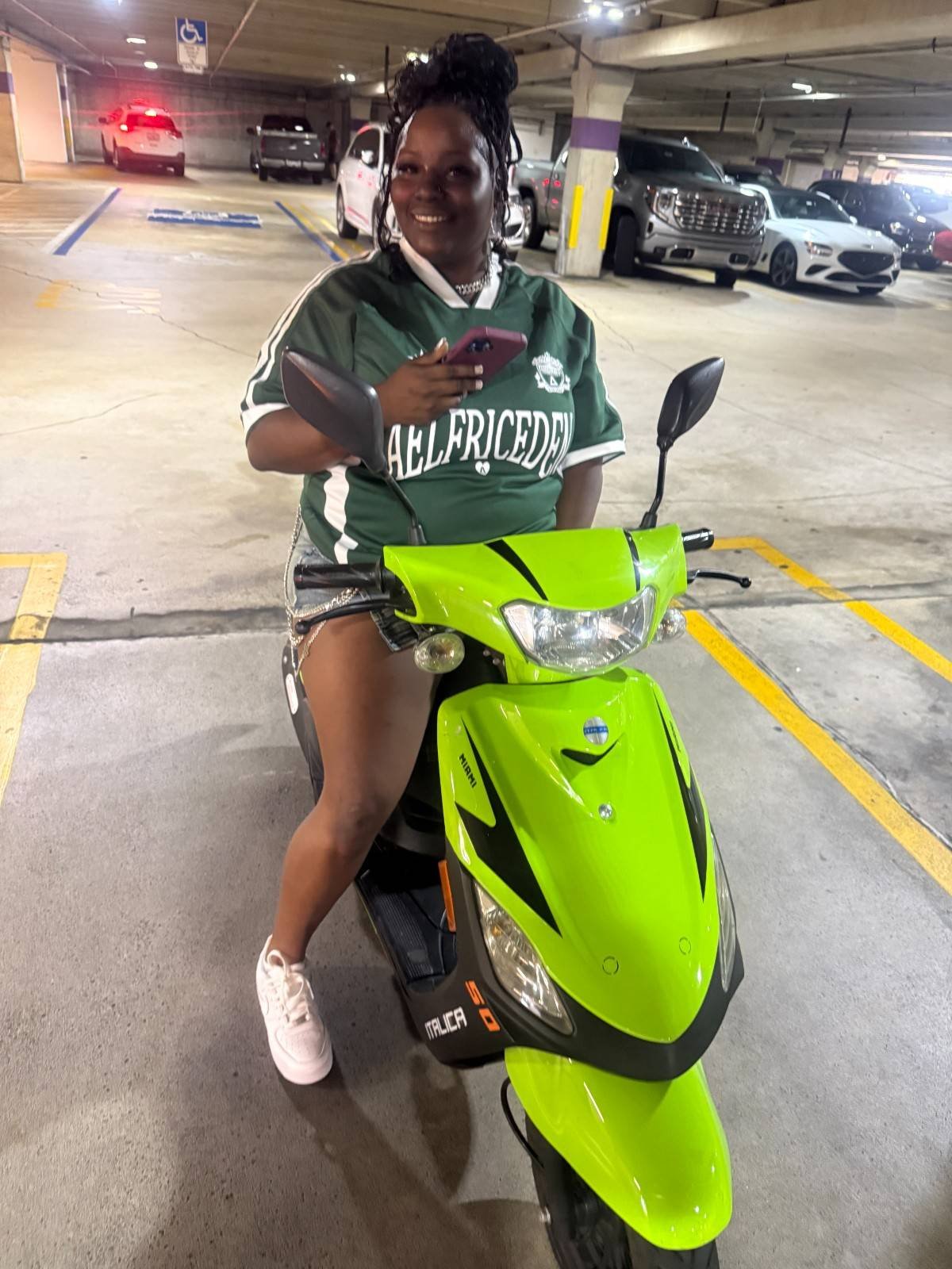 A woman in a green sports jersey and shorts sitting on a lime green scooter in a parking garage, holding a smartphone and smiling.