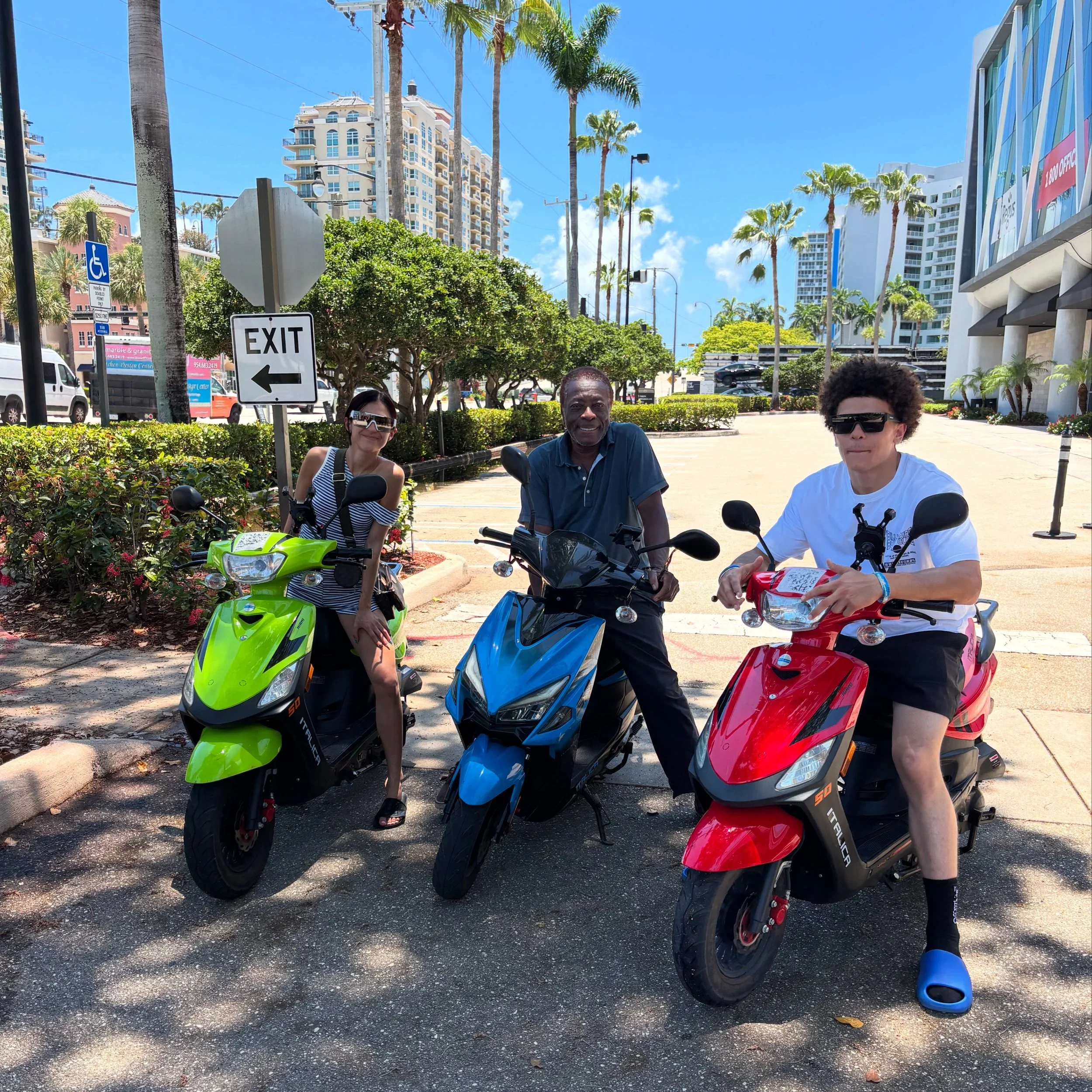 Three people sitting on scooters in an urban area with tall buildings, palm trees, street signs, and vehicles in the background on a sunny day.