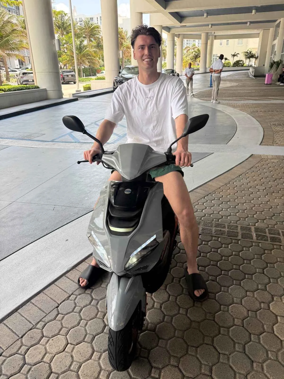 A man sitting on a silver scooter in a covered area outside a building, wearing a white t-shirt, shorts, and sandals, smiling at the camera.