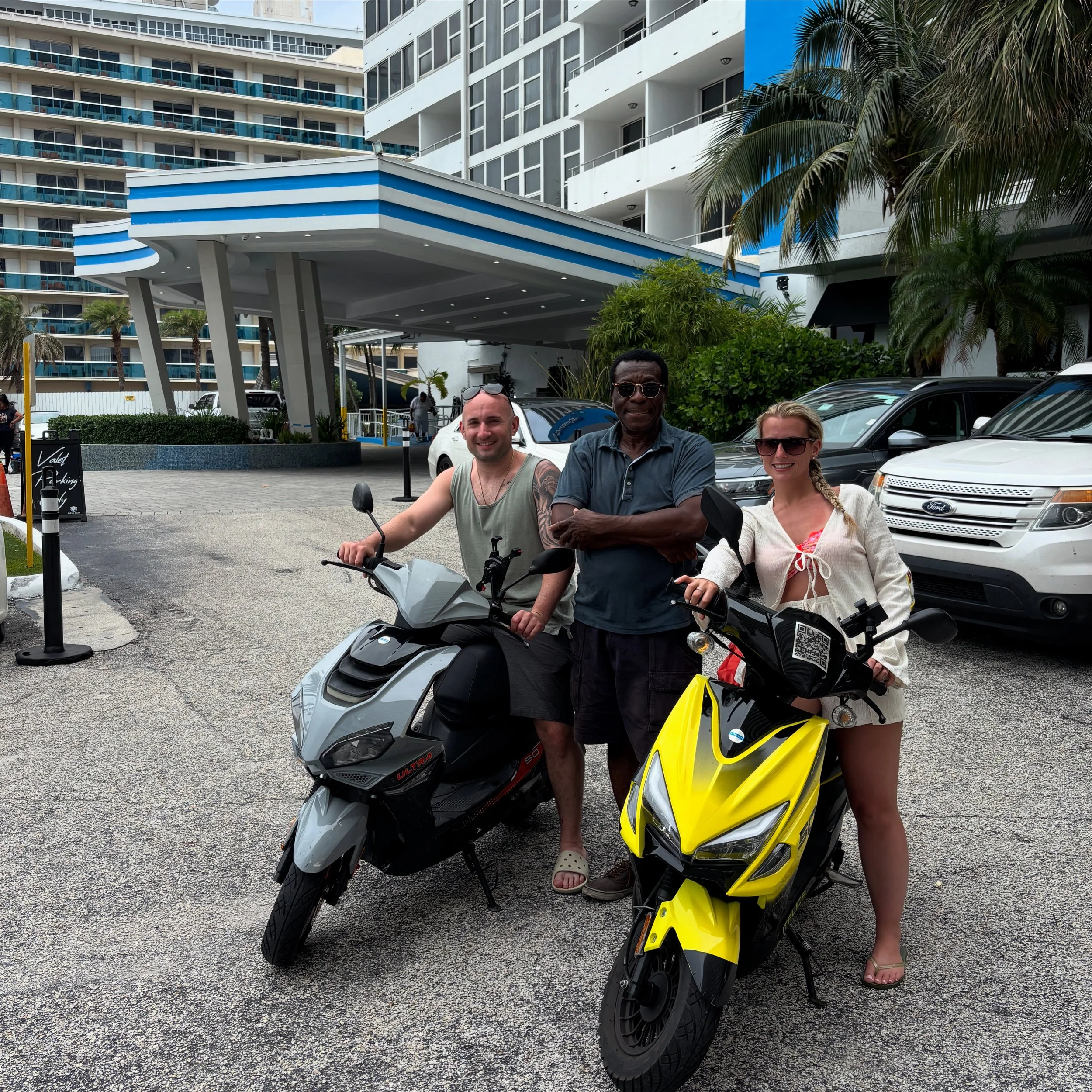 A group of three people standing next to scooters in front of a modern hotel building with balconies and palm trees, with one person sitting on a gray scooter and another on a yellow scooter.