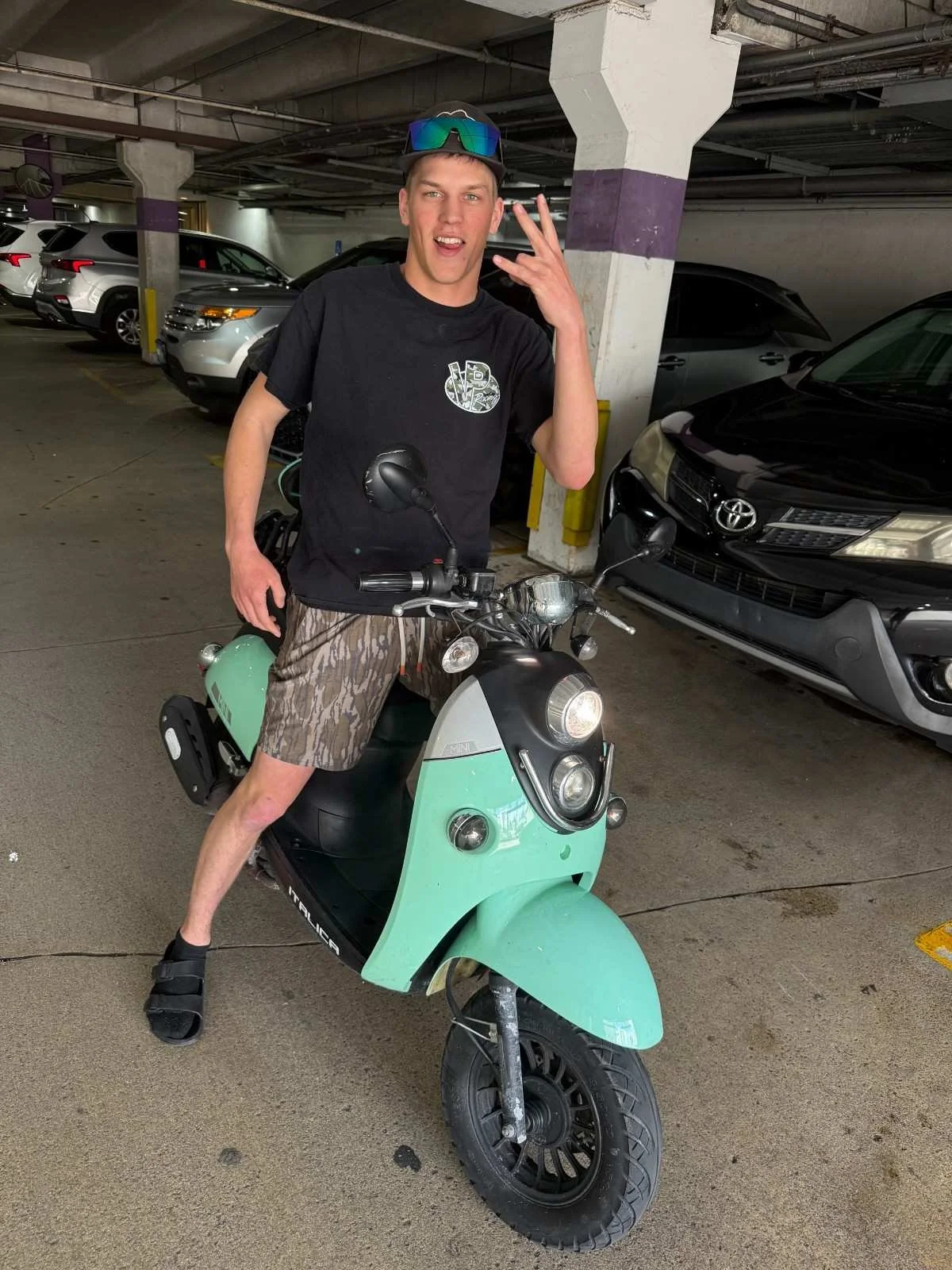 Young man making a gesture with his hand while sitting on a mint green scooter in a parking garage.