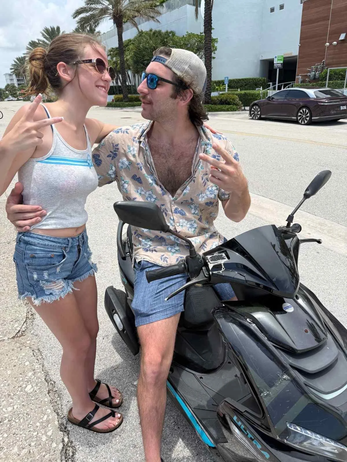 A young woman and a man sitting on a black scooter on a street. The woman is wearing sunglasses, a gray tank top, and denim shorts. The man is wearing a floral shirt, shorts, a cap, and sunglasses. Both are making peace signs and smiling at each othe