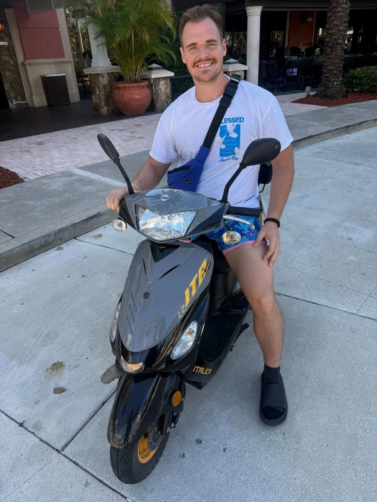 A smiling man wearing a white t-shirt, colorful shorts, black slides, and a black wristwatch, sitting on a black Italica scooters scooter, parked on a sidewalk in front of a building with palm trees and potted plants.