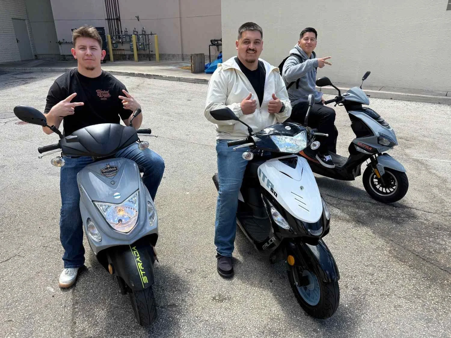 Three men sitting on scooters in a parking lot, posing for the camera, with a plain wall and some industrial pipes in the background.