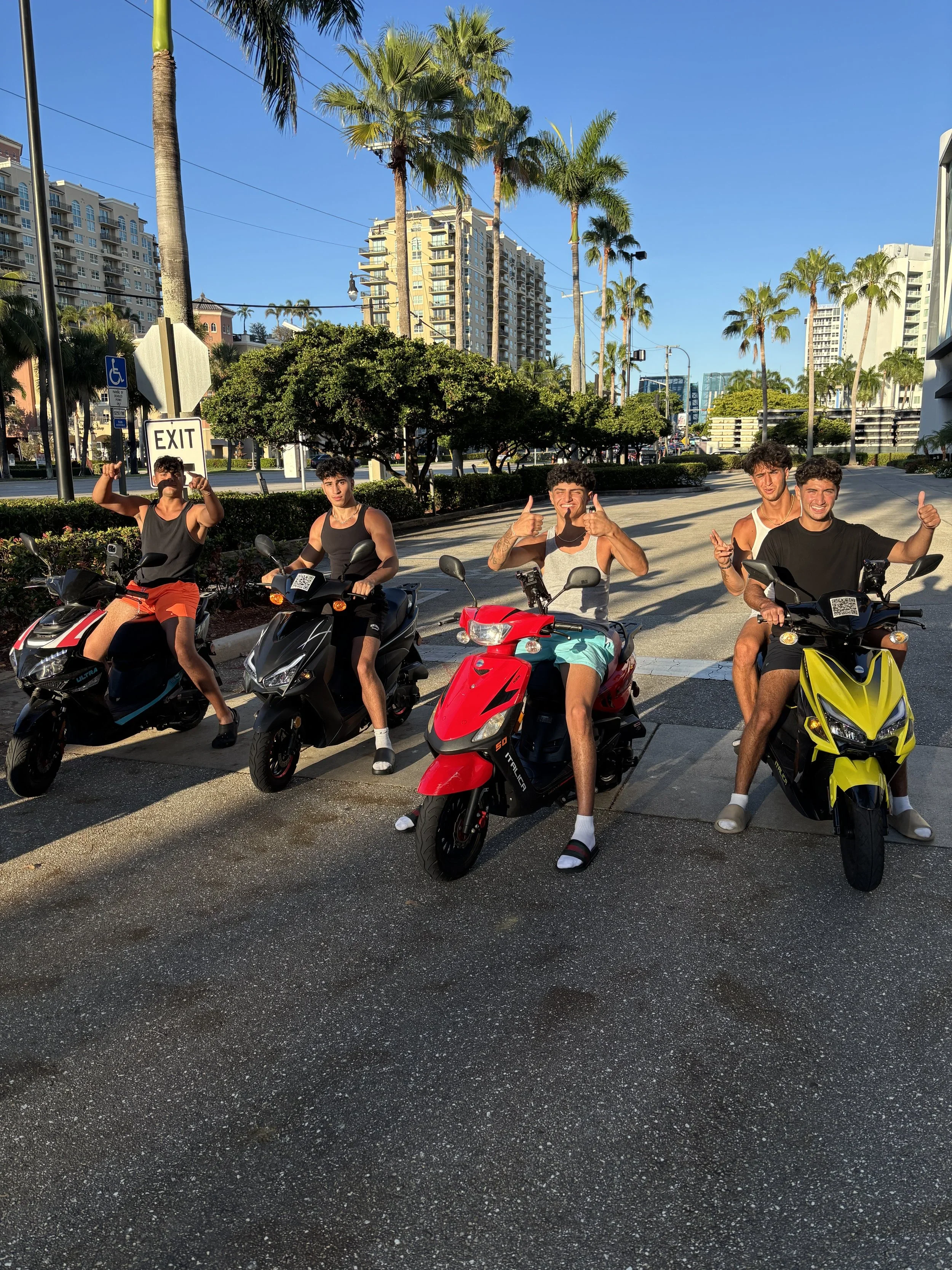 Five young men on scooters, posing for a photo on a sunny city street with modern buildings and tall palm trees in the background. They are smiling, with some giving thumbs-up and confident gestures.