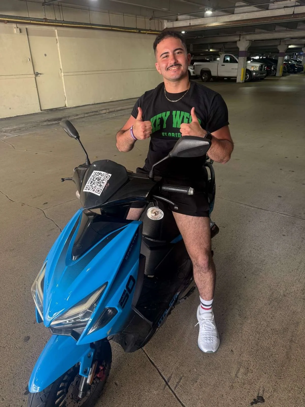 Man sitting on a blue electric scooter in an underground parking garage, smiling and giving two thumbs up. He is wearing a black t-shirt with green text, black shorts, white sneakers, and striped socks.