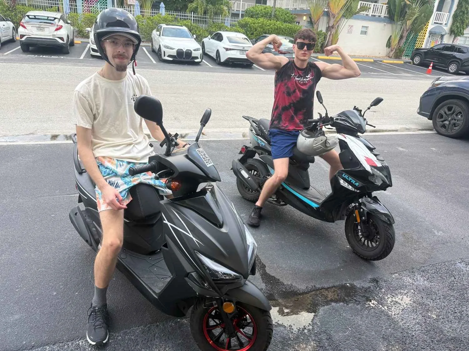 Two young men on scooters in a parking lot, one wearing a helmet and casual clothes, the other flexing muscles and wearing sunglasses and a sleeveless shirt.