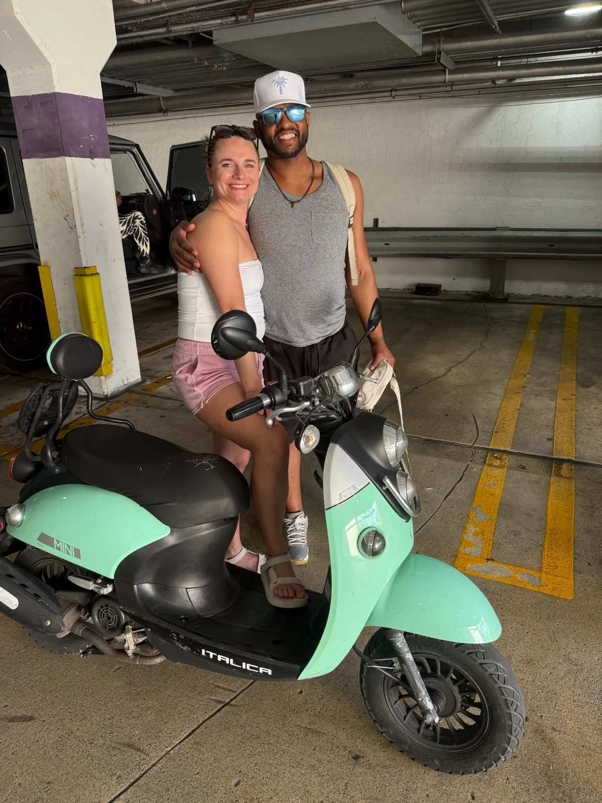 A woman sitting on a mint green scooter in an underground parking garage, embracing a smiling man with a beard. The woman is wearing a white tank top and pink shorts, while the man is wearing sunglasses, a gray sleeveless shirt, and a white cap. He h