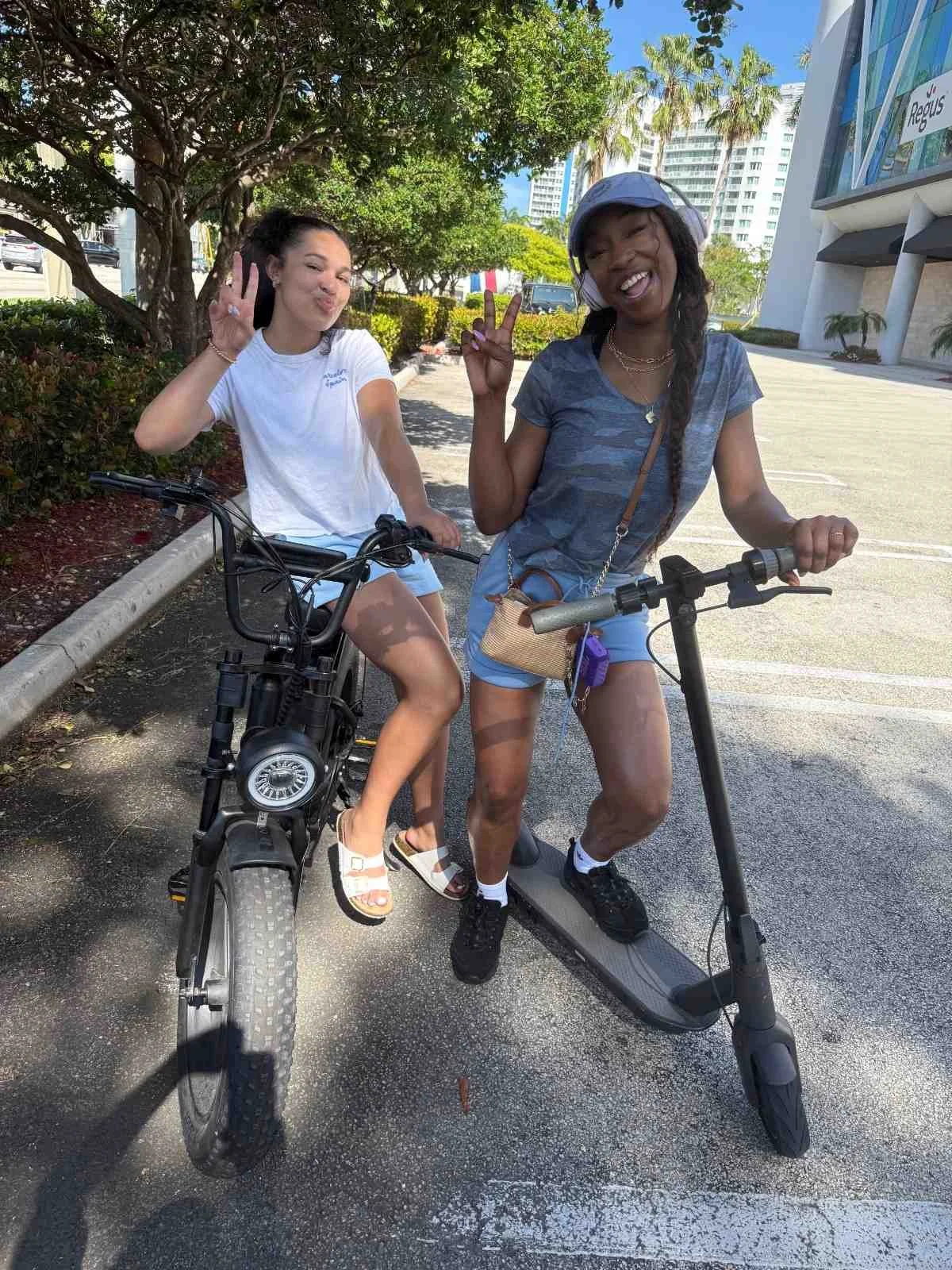 Two women, one on an electric scooter and the other on a bicycle, smiling and making peace signs outdoors on a sunny day with trees and modern buildings in the background.