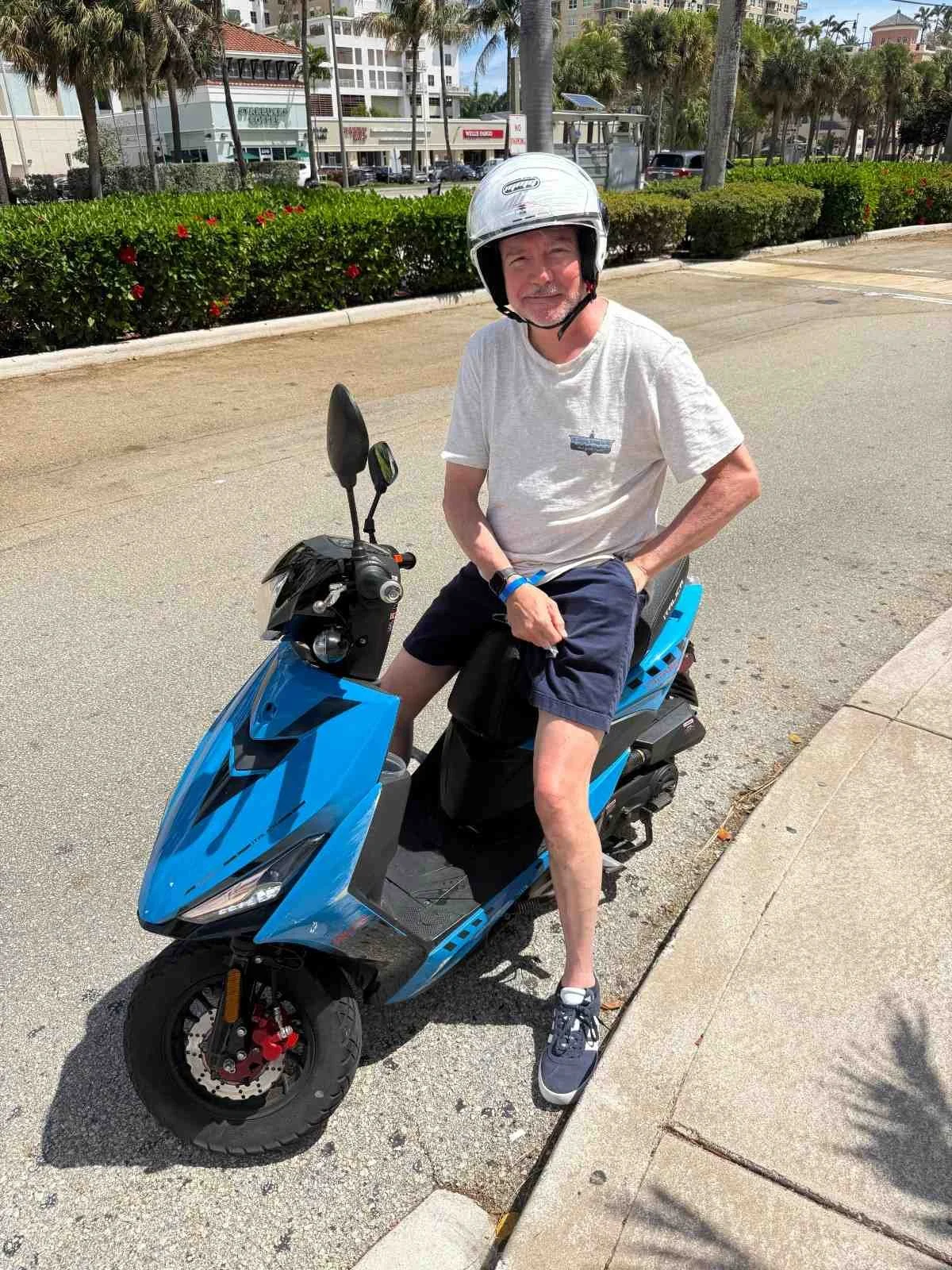 An older man wearing a white helmet, white t-shirt, dark shorts, and sneakers sitting on a blue electric scooter on the sidewalk amid a sunny day in an urban area with palm trees and modern buildings in the background.