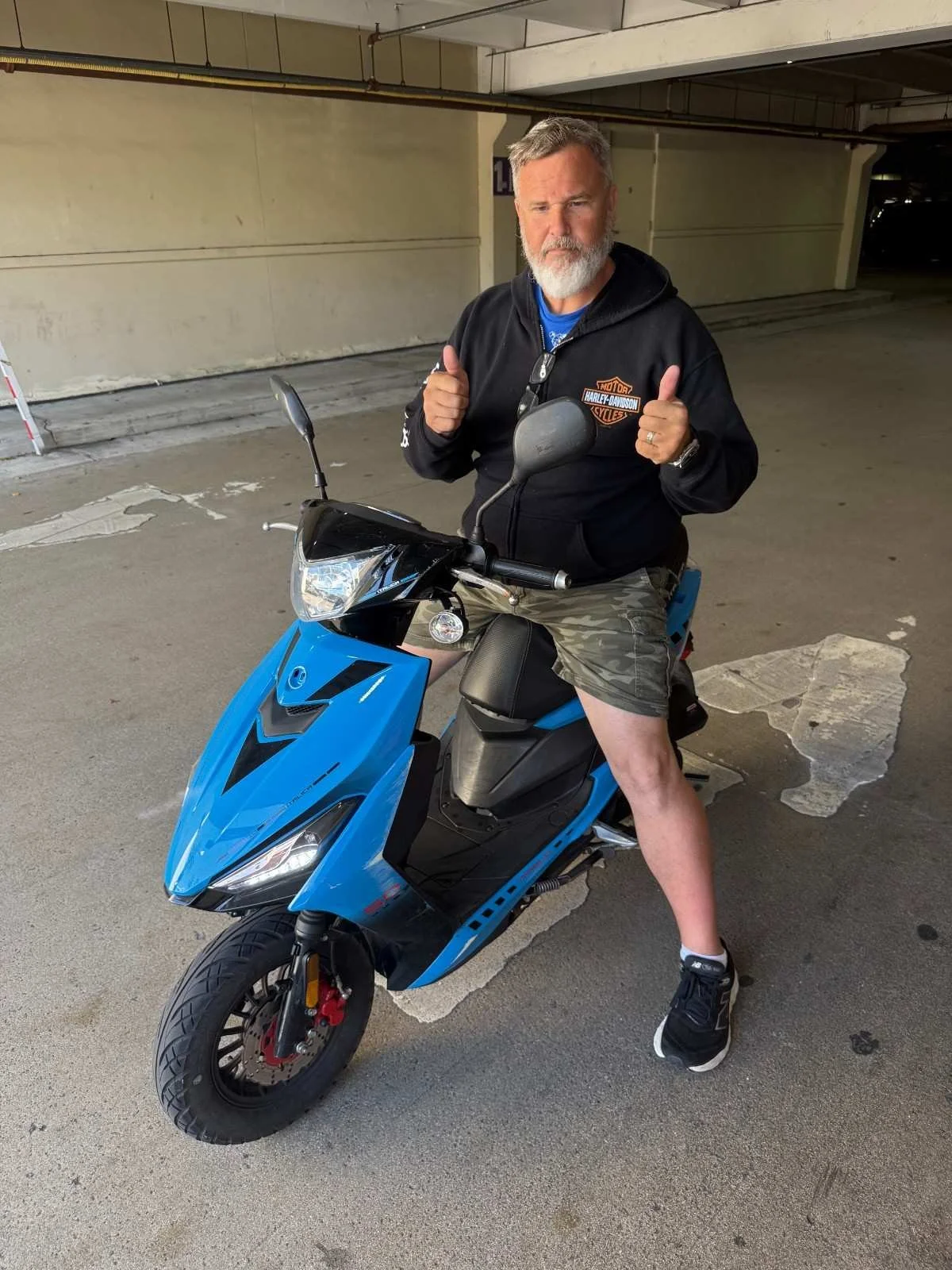 A man with gray hair and beard, wearing a black Harley Davidson hoodie, camo shorts, and sneakers, sitting on a blue scooter in a parking garage, giving two thumbs up.