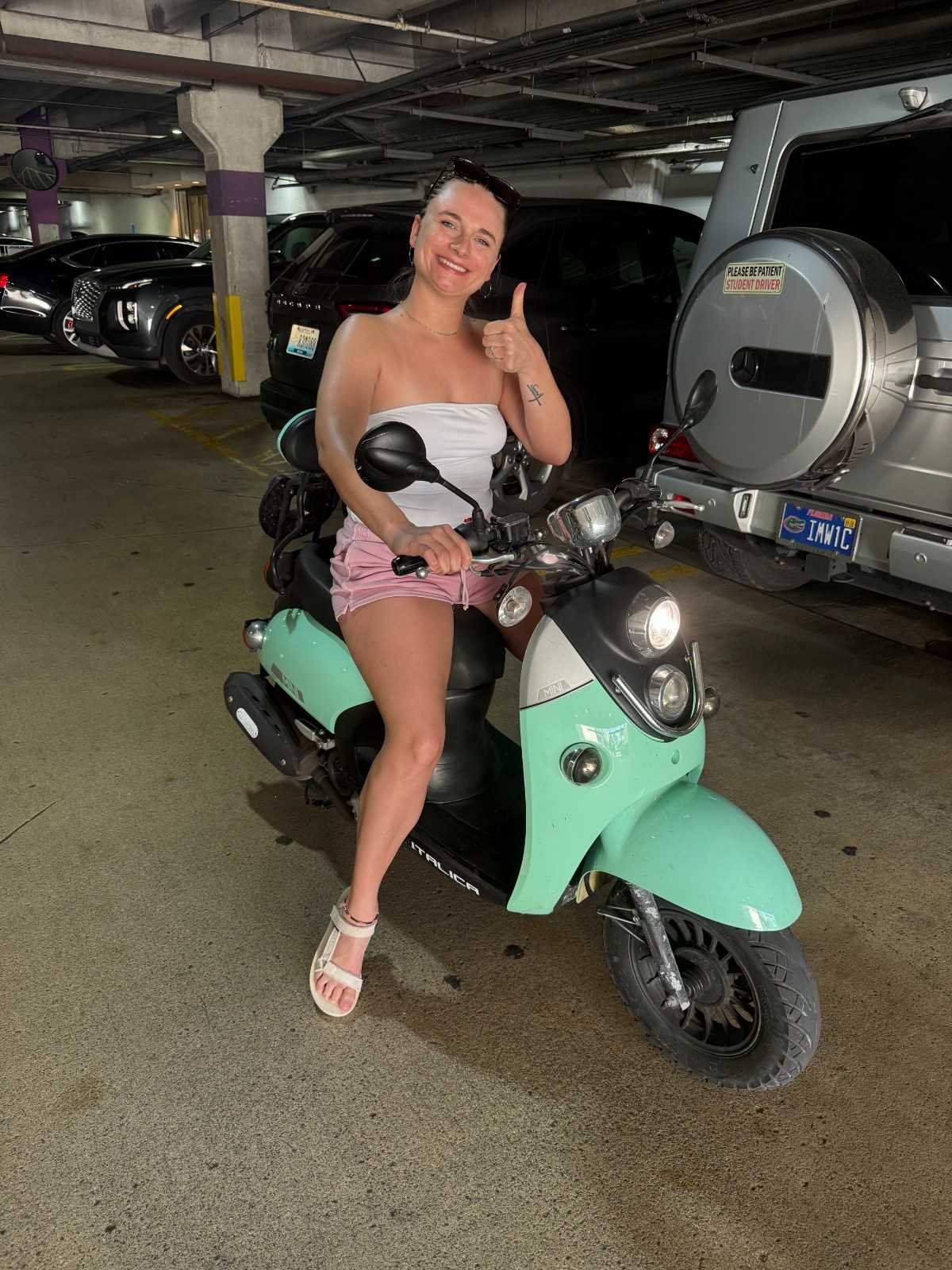 A woman in a strapless white top and pink shorts giving a thumbs up while sitting on a mint green electric scooter in a parking garage.
