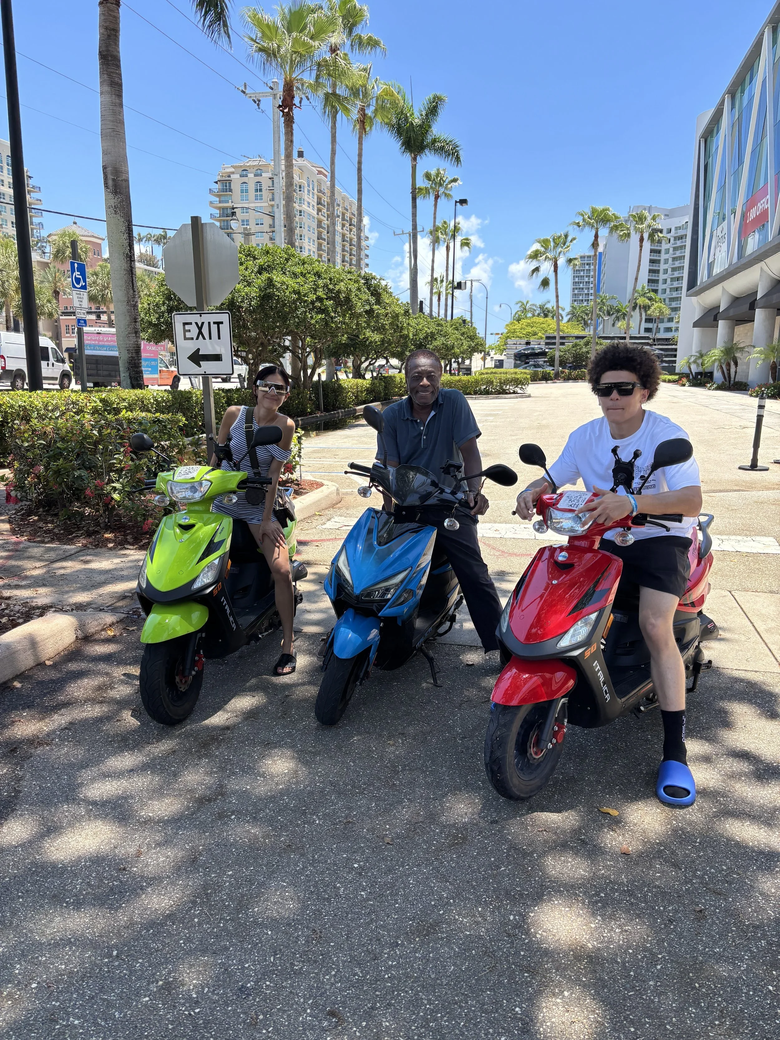 Three people sitting on colorful scooters in a sunny outdoor urban area with palm trees and modern buildings in the background.